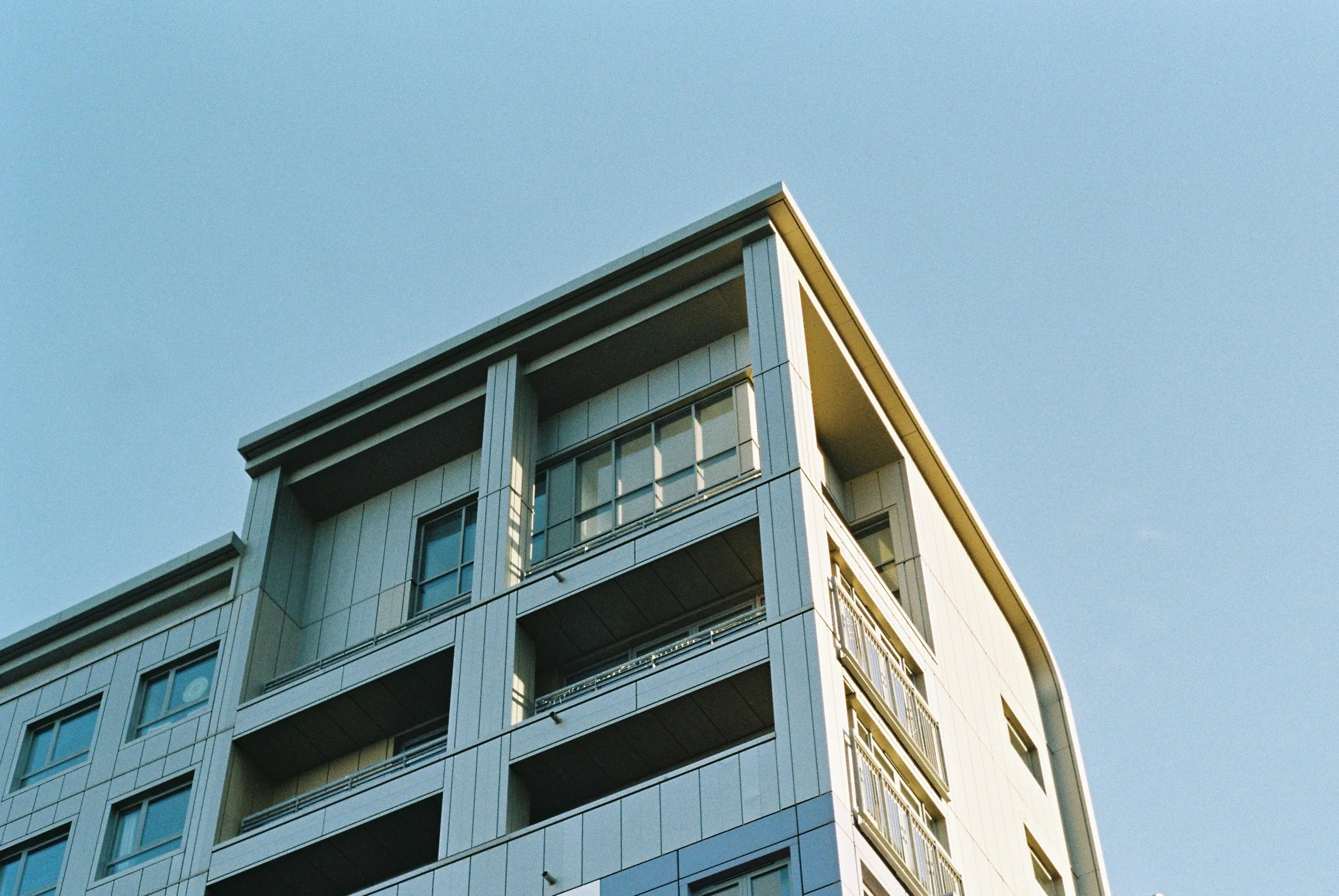 A modern multi-story building with balconies and large windows, set against a clear blue sky.