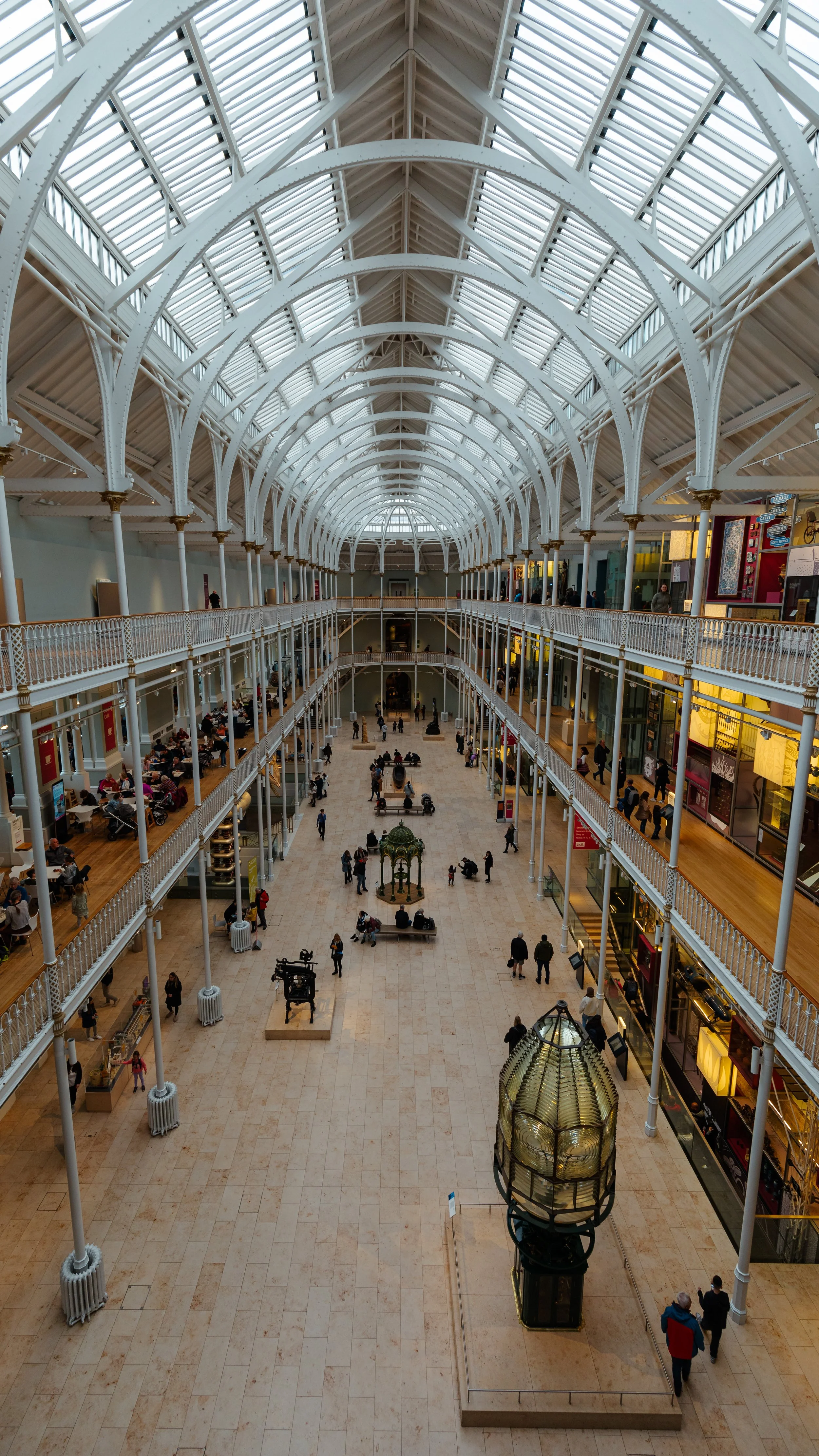 Interior view of a large shopping mall with glass ceiling, multiple floors with stores, and many people walking and sitting in the open space.