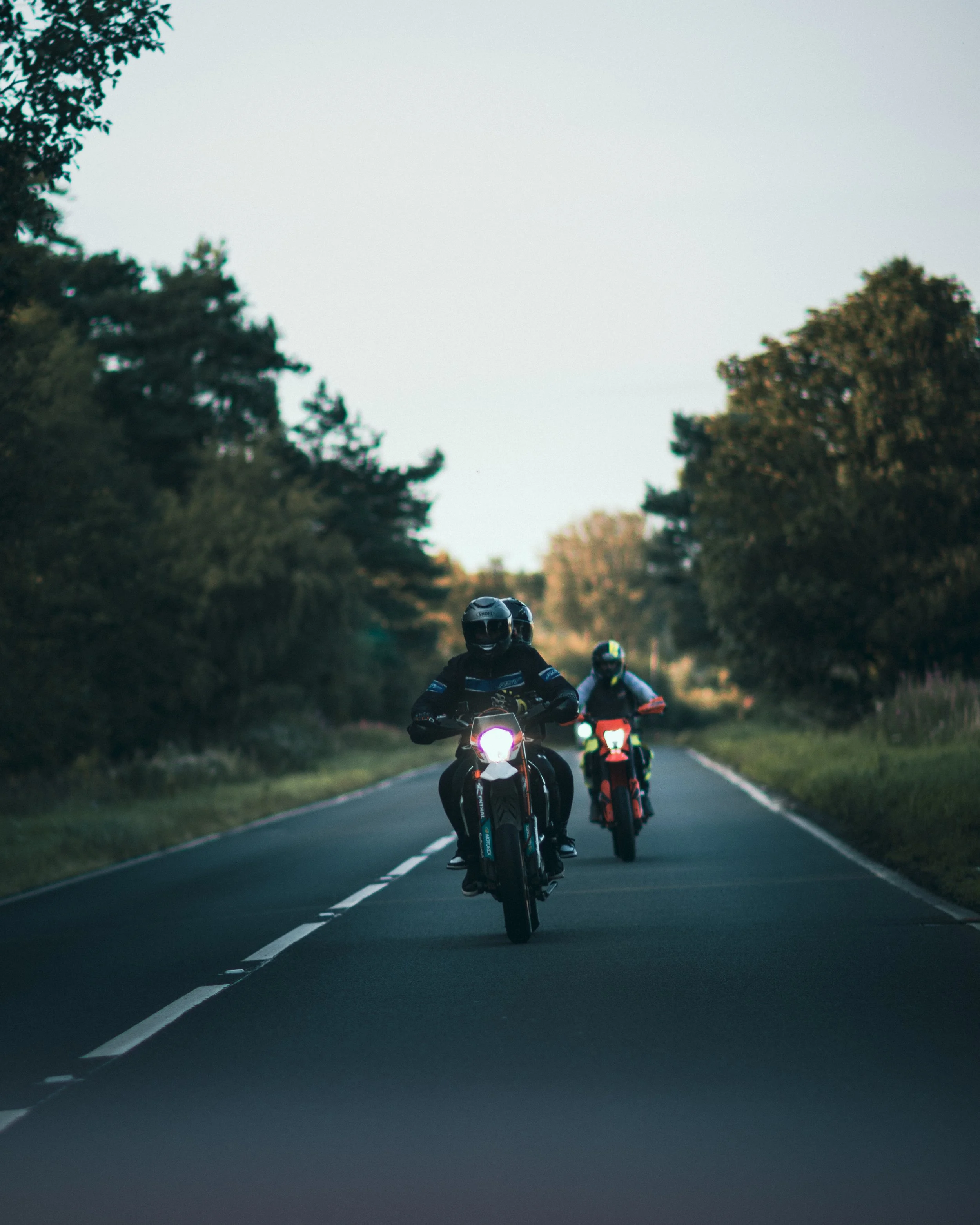 Three motorcyclists riding on a two-lane road surrounded by trees, with the lead rider wearing a black helmet and black gear, and the others following behind.