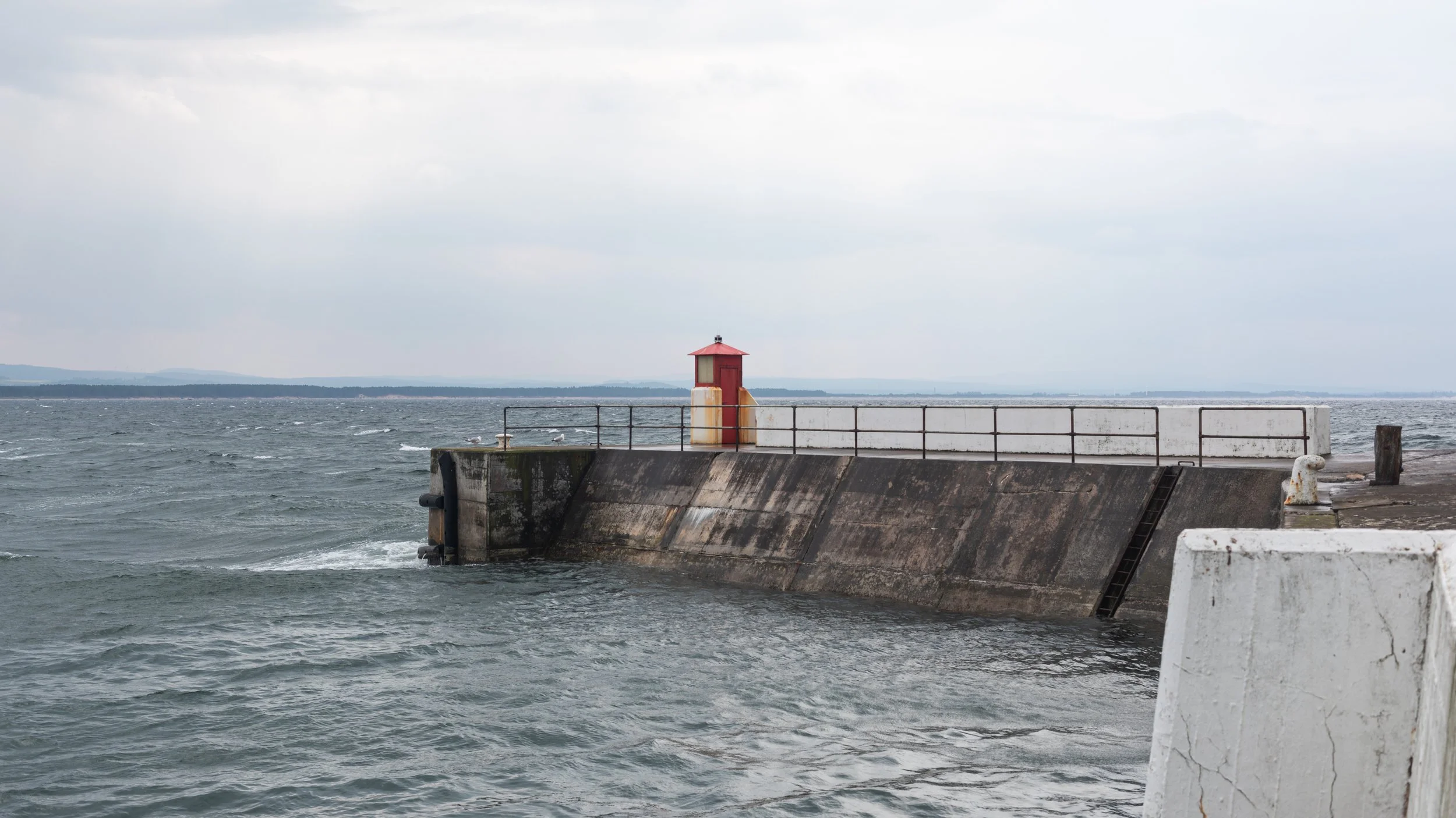 A concrete pier extending into the sea with a small red lighthouse, metal railing, and seagulls on the water, under a cloudy sky.