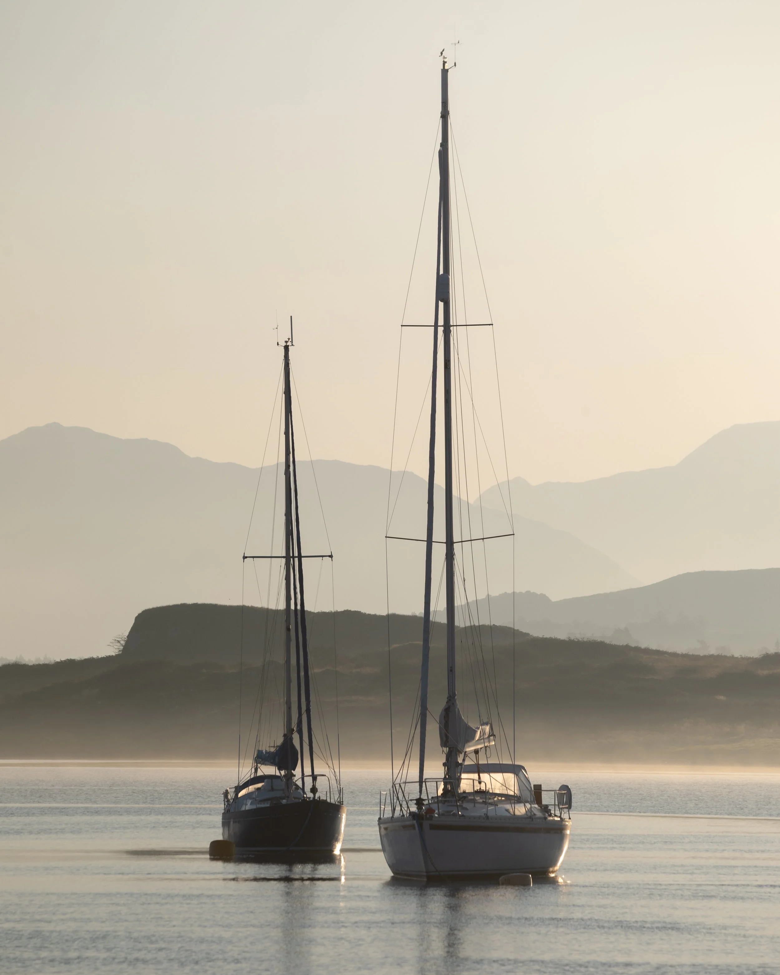 Two sailboats floating on calm water with misty mountains in the background, during sunset or sunrise.