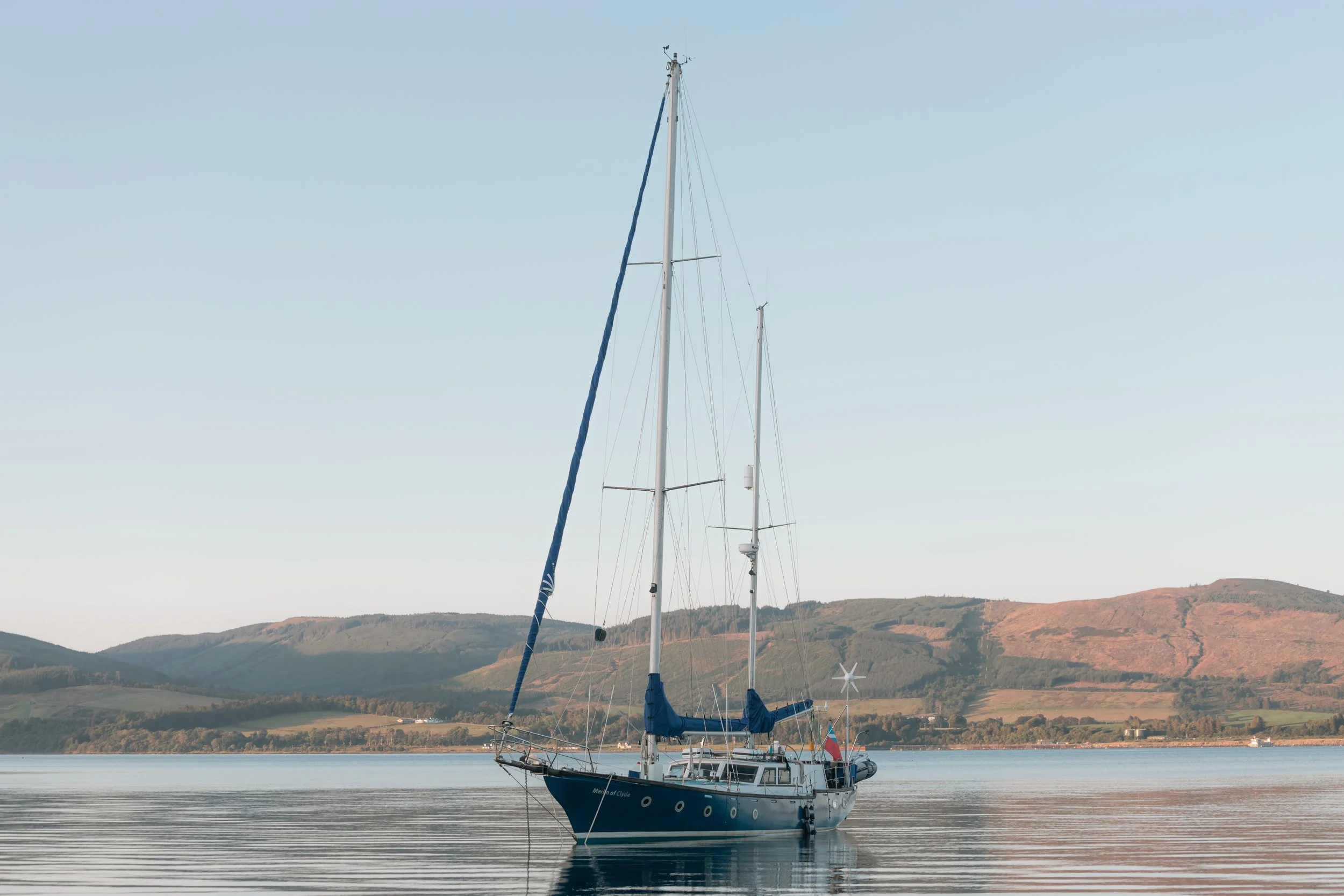 Sailboat named 'Merlin of Clyne' floating on calm water with hills in the background under a clear sky.