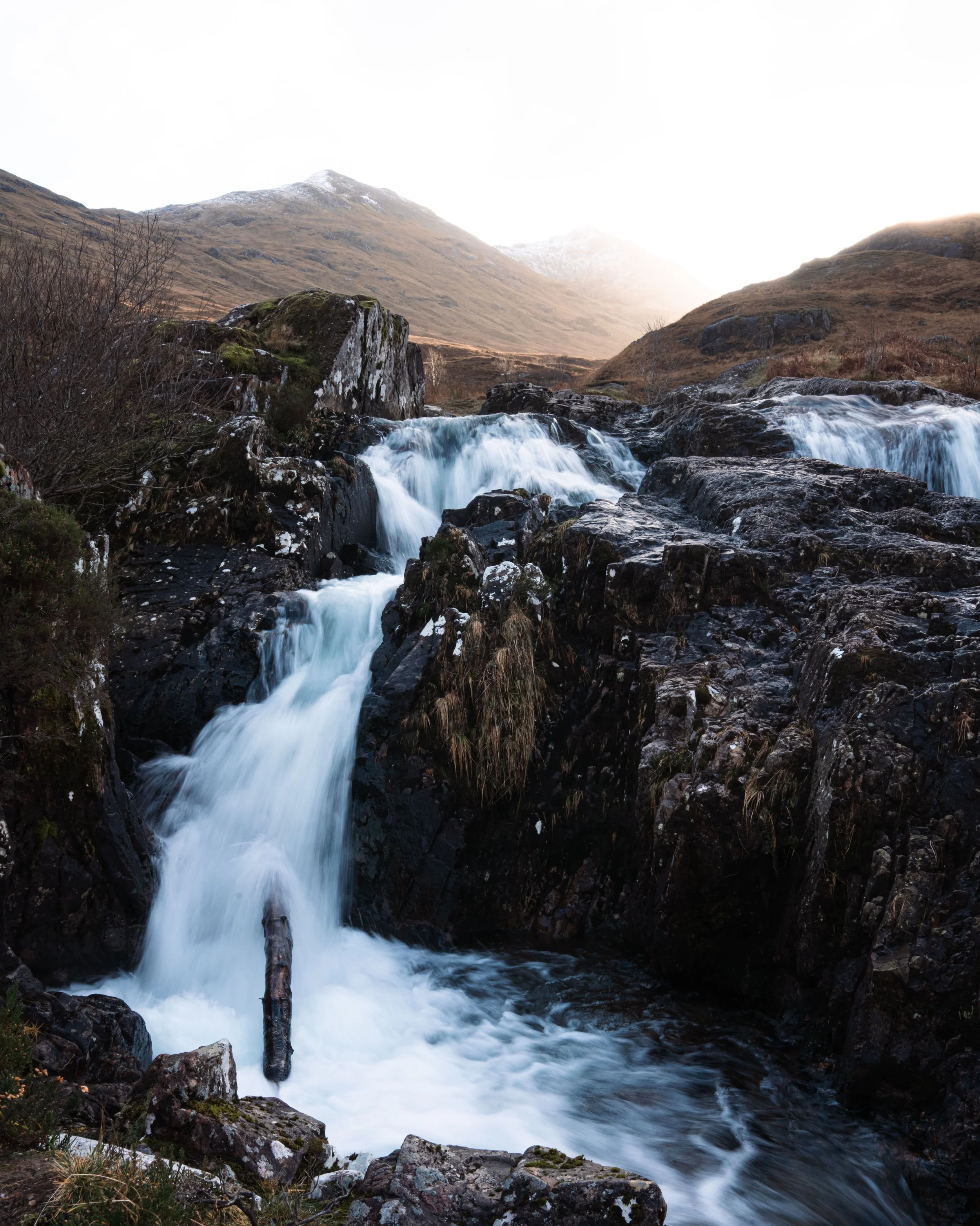A small waterfall flowing over rocks in a mountain valley with hills and a snow-capped peak in the background.