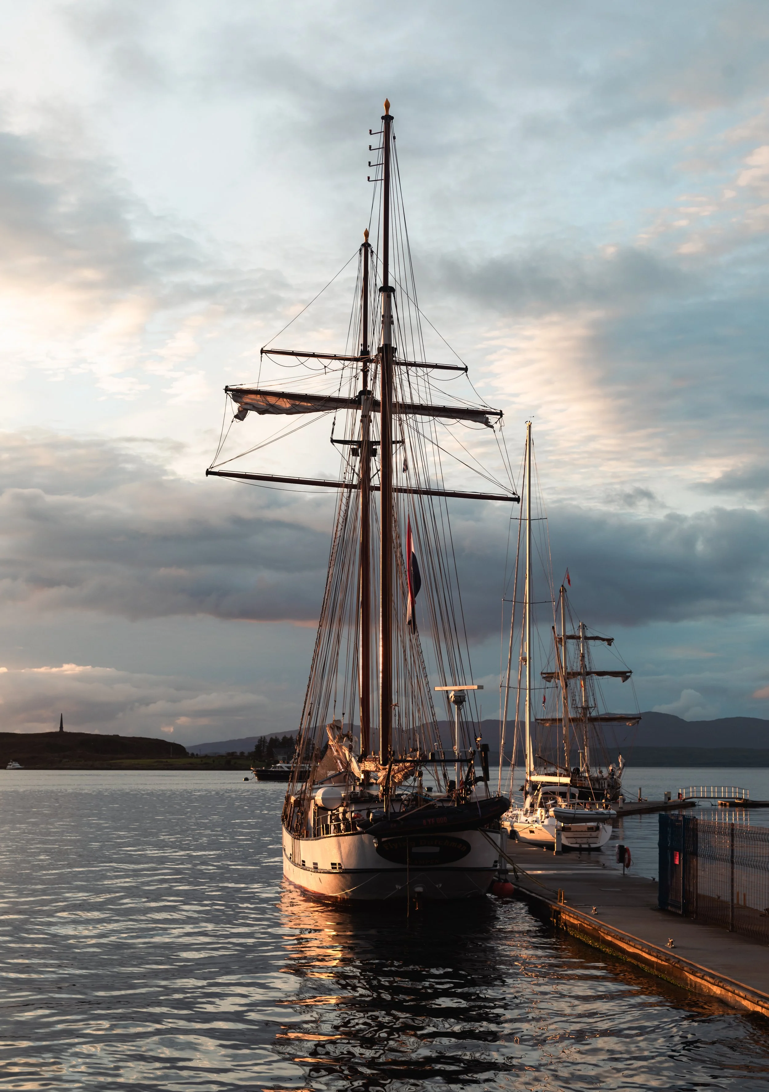 Sailboats docked at a marina during sunset with hills and water in the background.