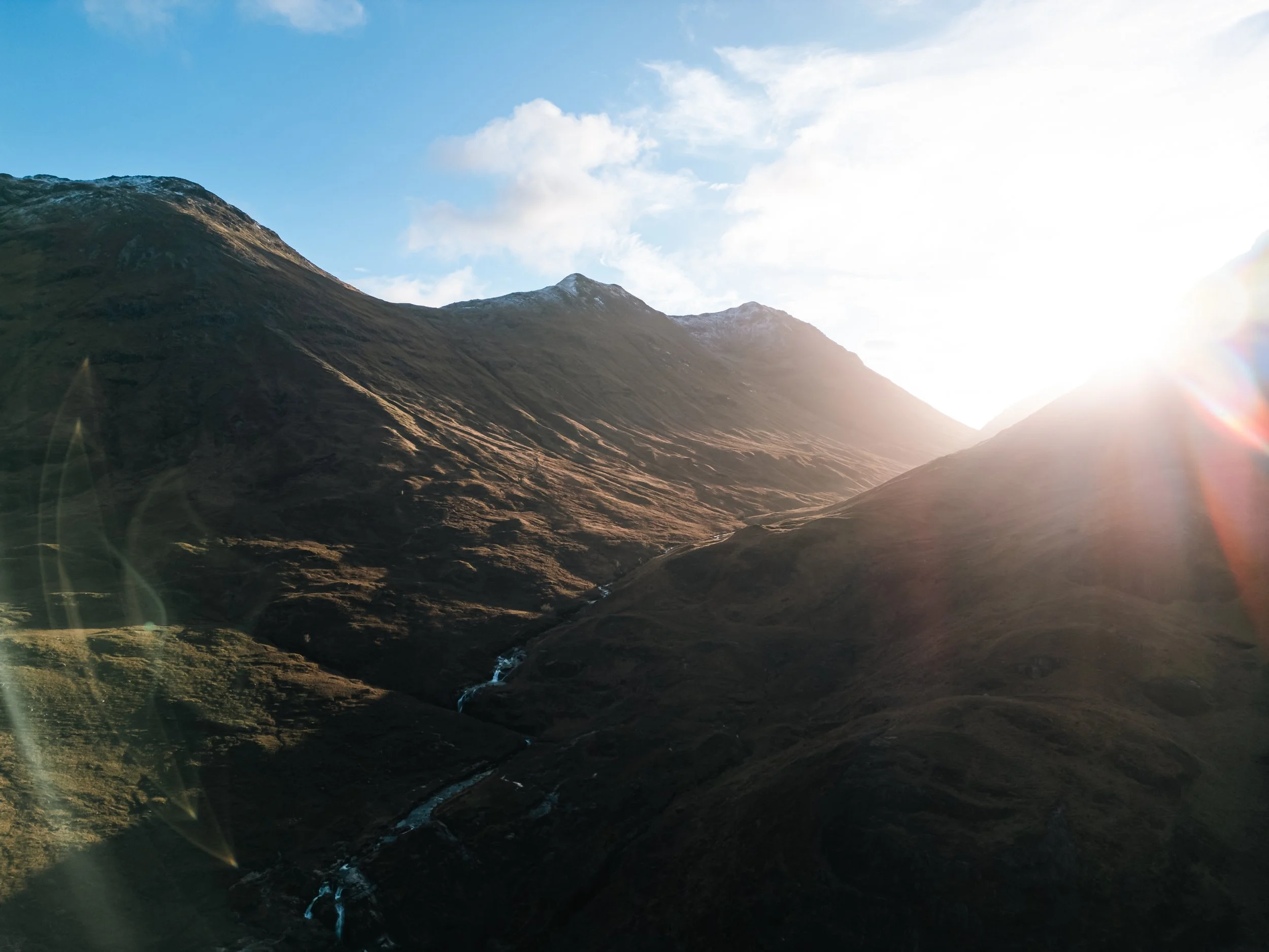 Sunset over mountains with a valley and a small river, bright sunlight creating a lens flare effect.