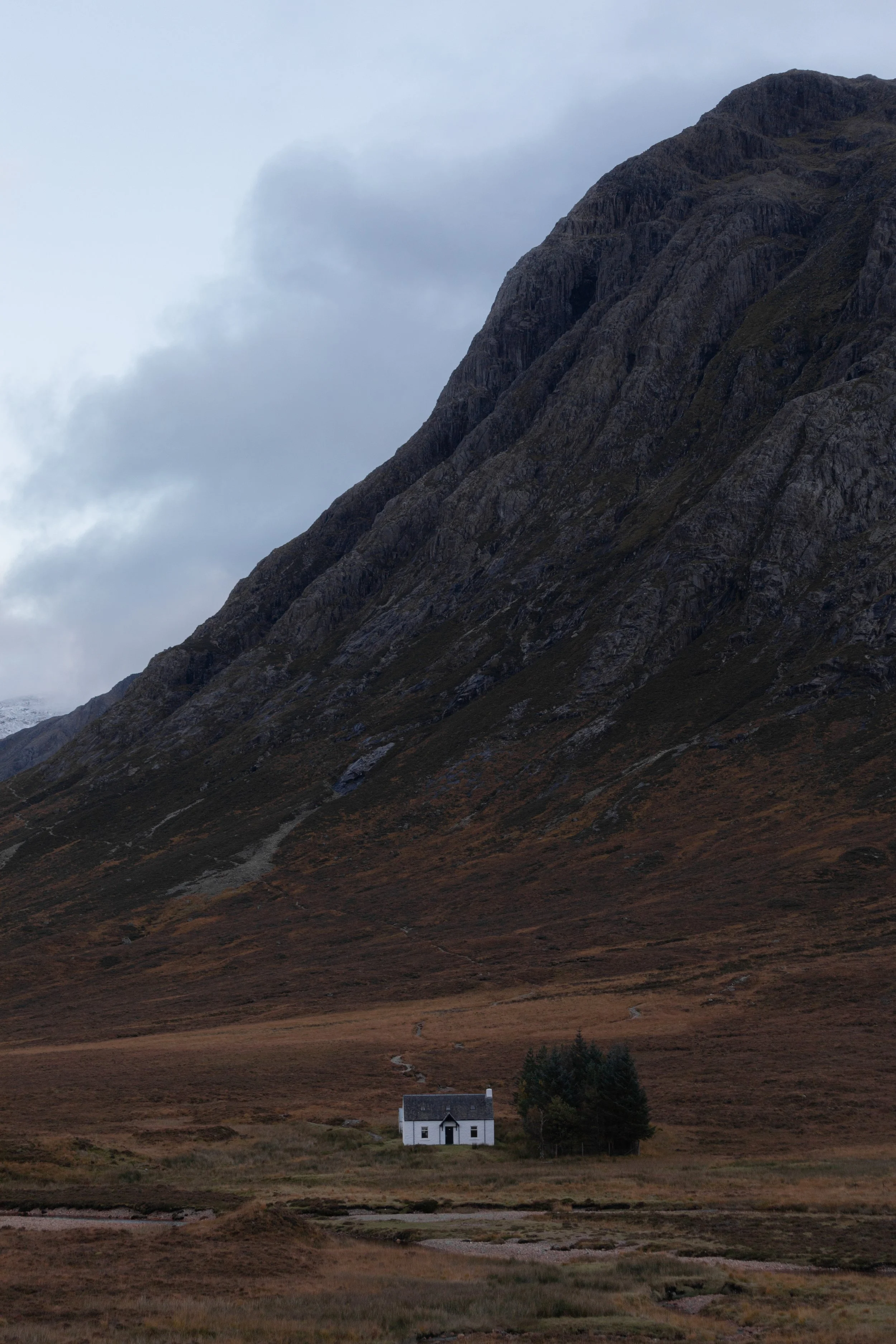 A small white house with dark roof and trim located in a vast open field at the base of towering mountain slopes, under a cloudy sky.