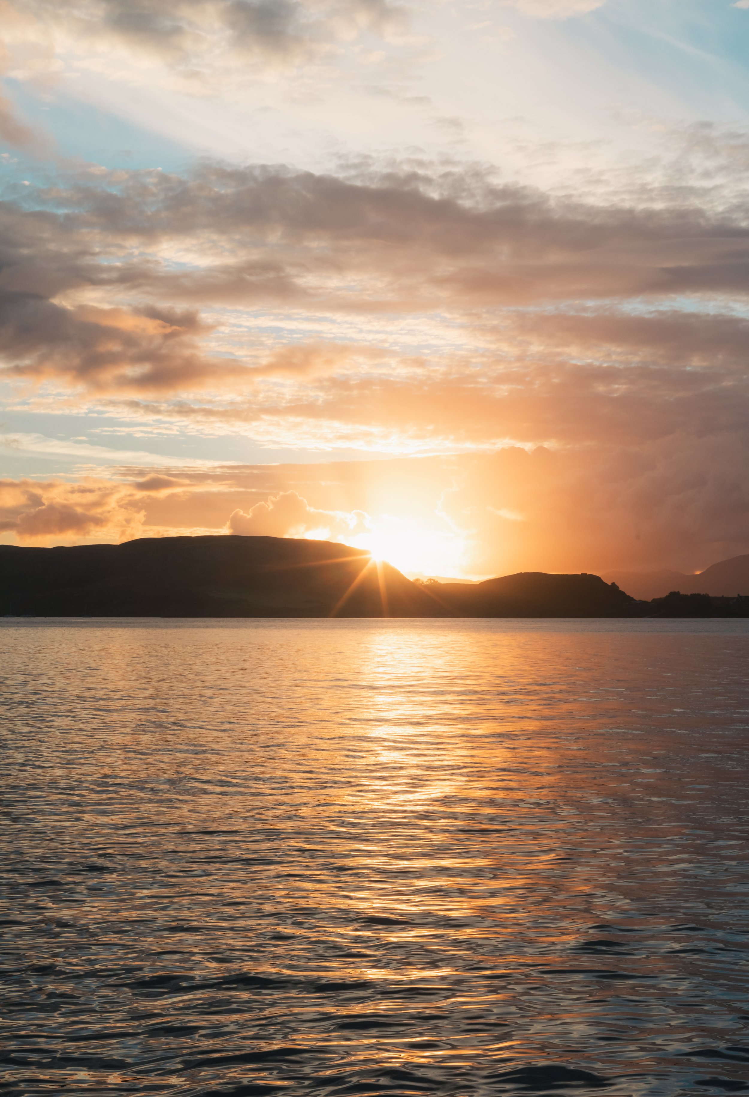 Sunset over a body of water with hills in the distance, partly cloudy sky with orange and blue hues.