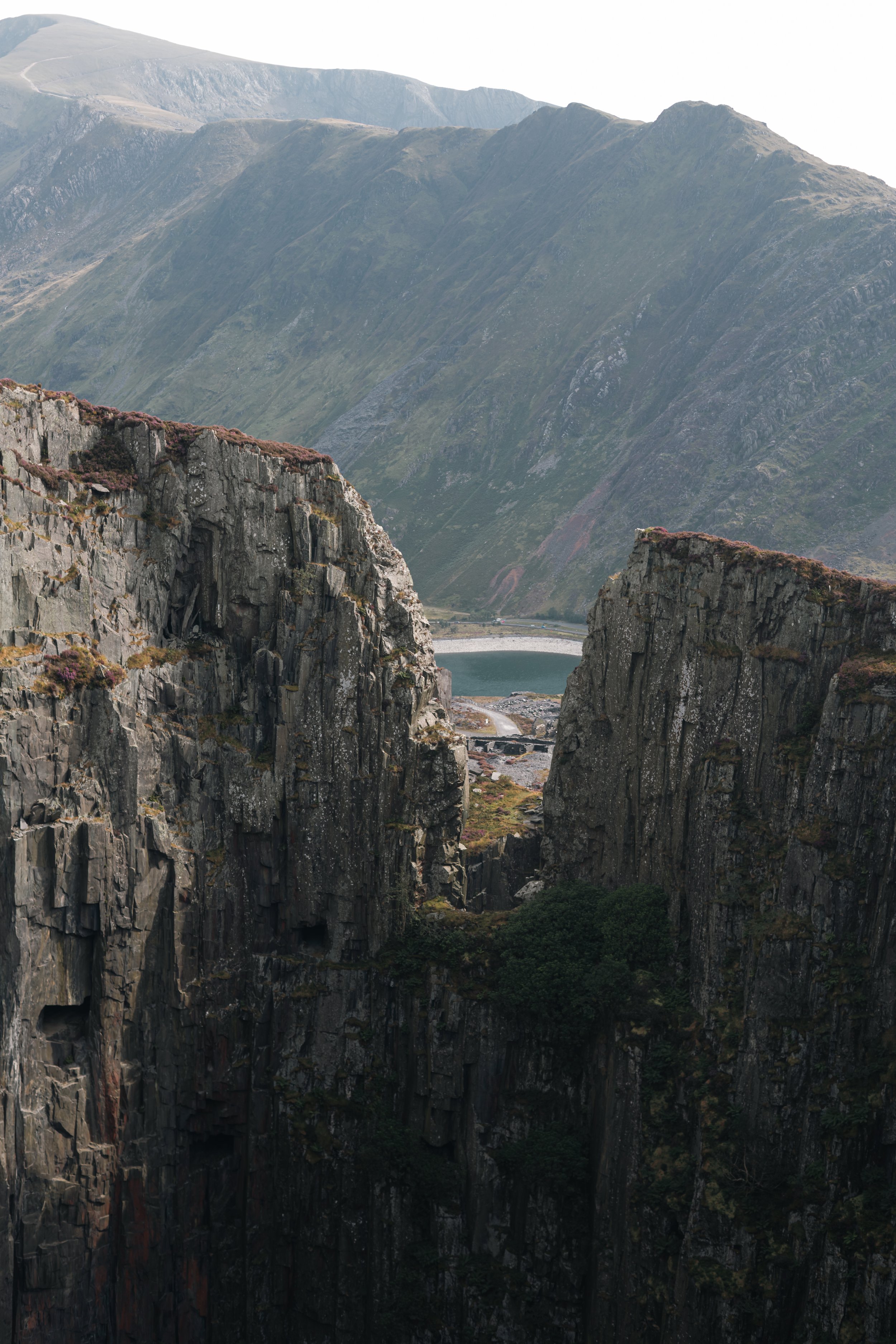 A scenic view of large, rugged granite cliffs with patches of grass and low vegetation, overlooking a lake at the base of towering mountains in the background.
