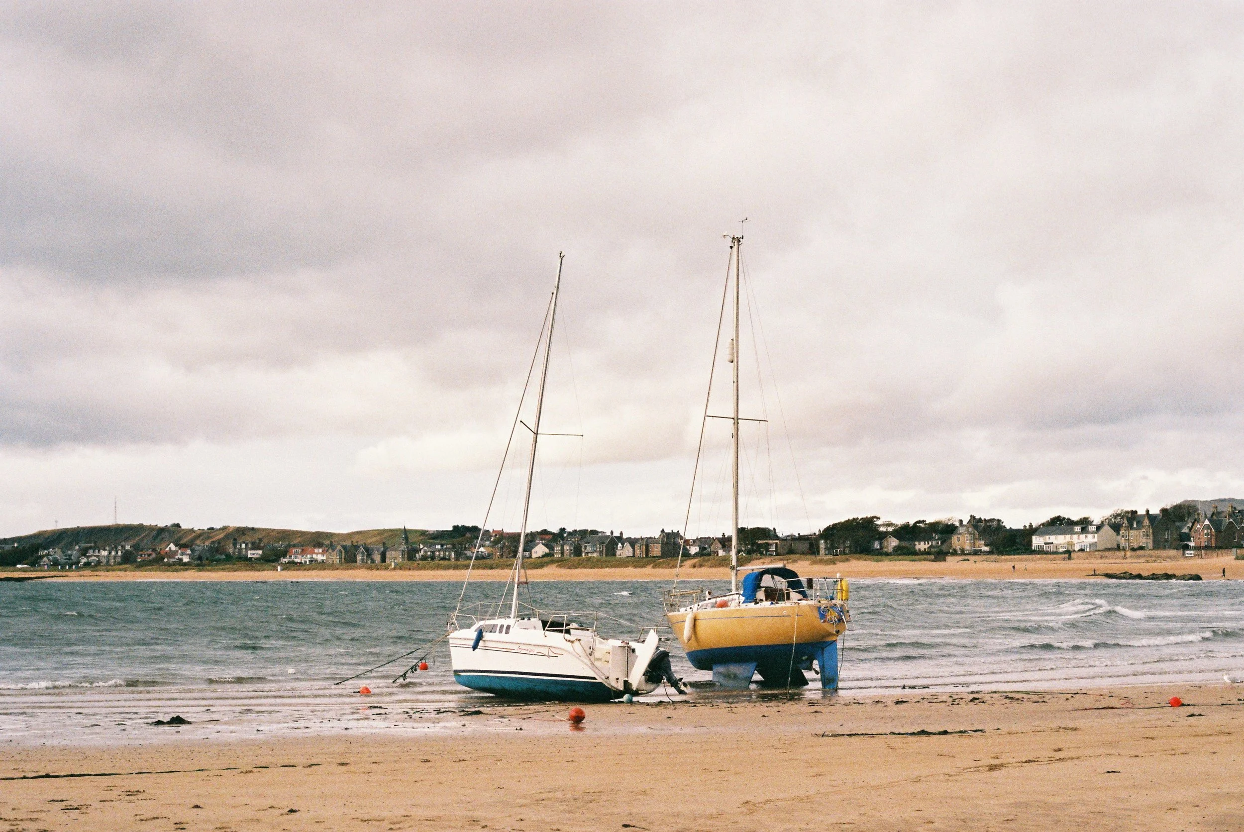 Two sailboats are grounded on a sandy beach with a town visible in the background under a cloudy sky.