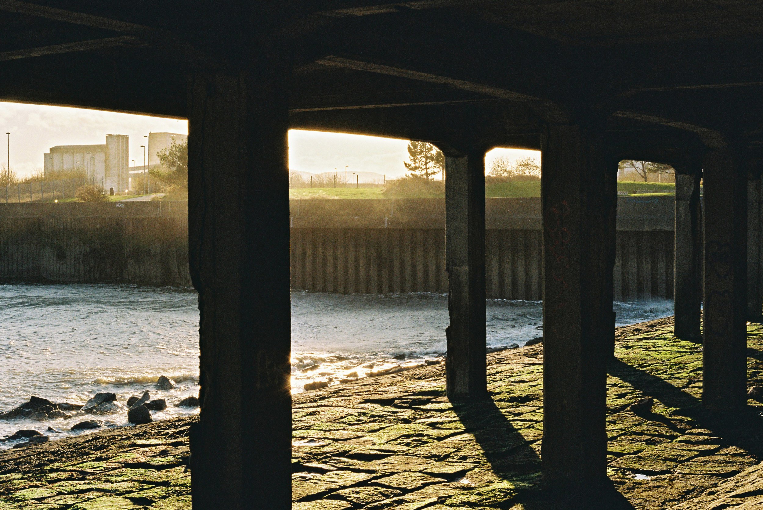 Under a bridge over a river, with sunlight shining through. The underside of the bridge supports is visible, and there are industrial buildings and trees in the background.