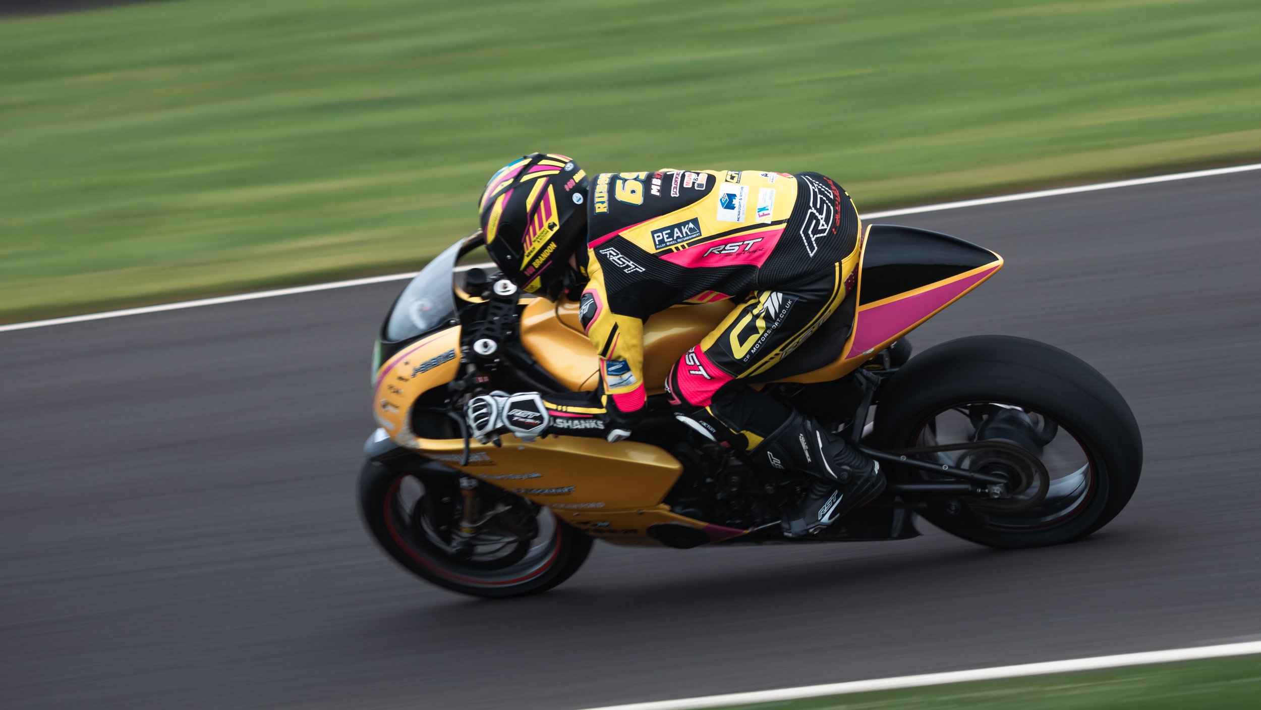 Motorcyclist in racing gear riding a yellow and black sport motorcycle on a racetrack with green grass in the background.