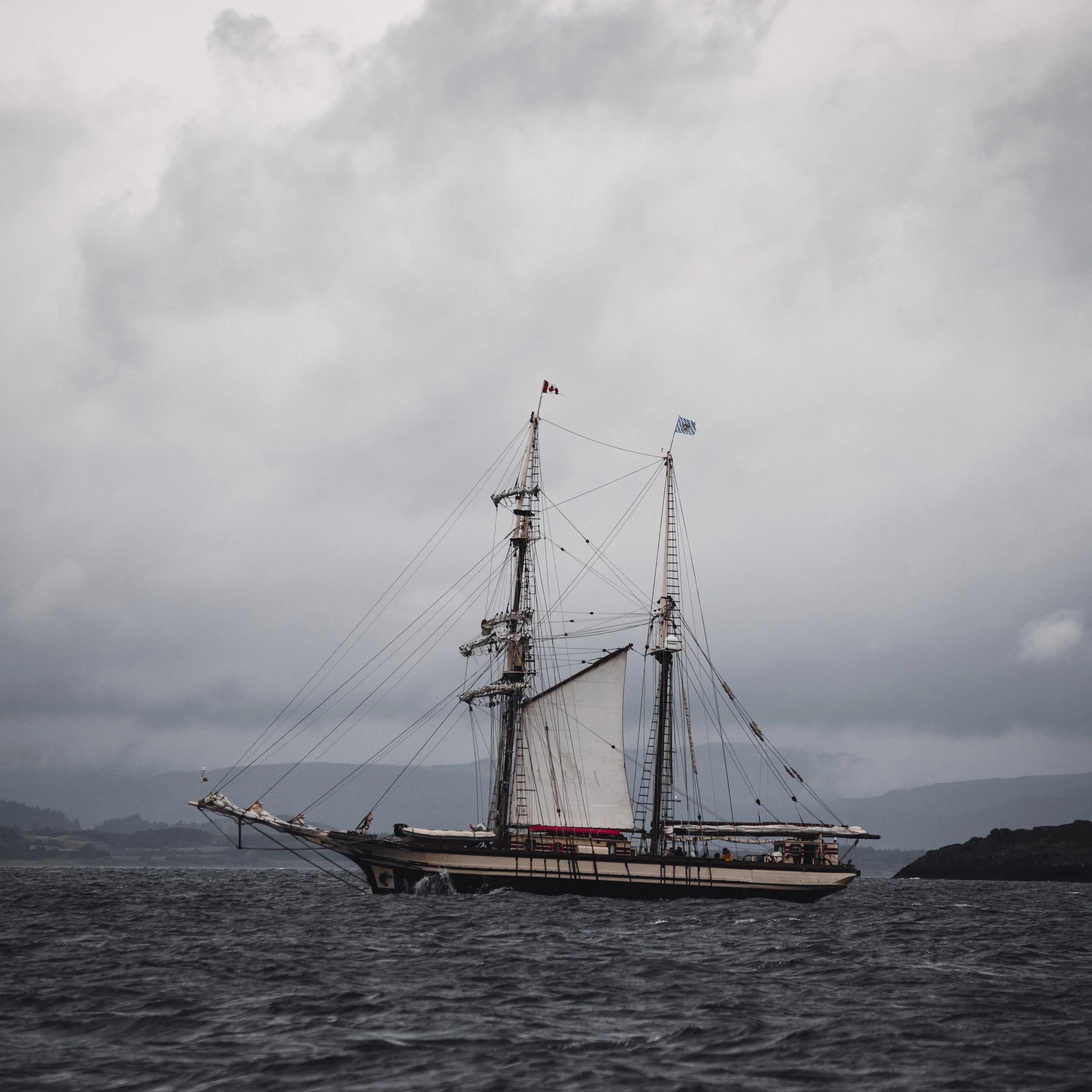 A large sailing ship with multiple masts, rigging, and sails, sailing on dark water under a cloudy gray sky, with flags flying on the masts.