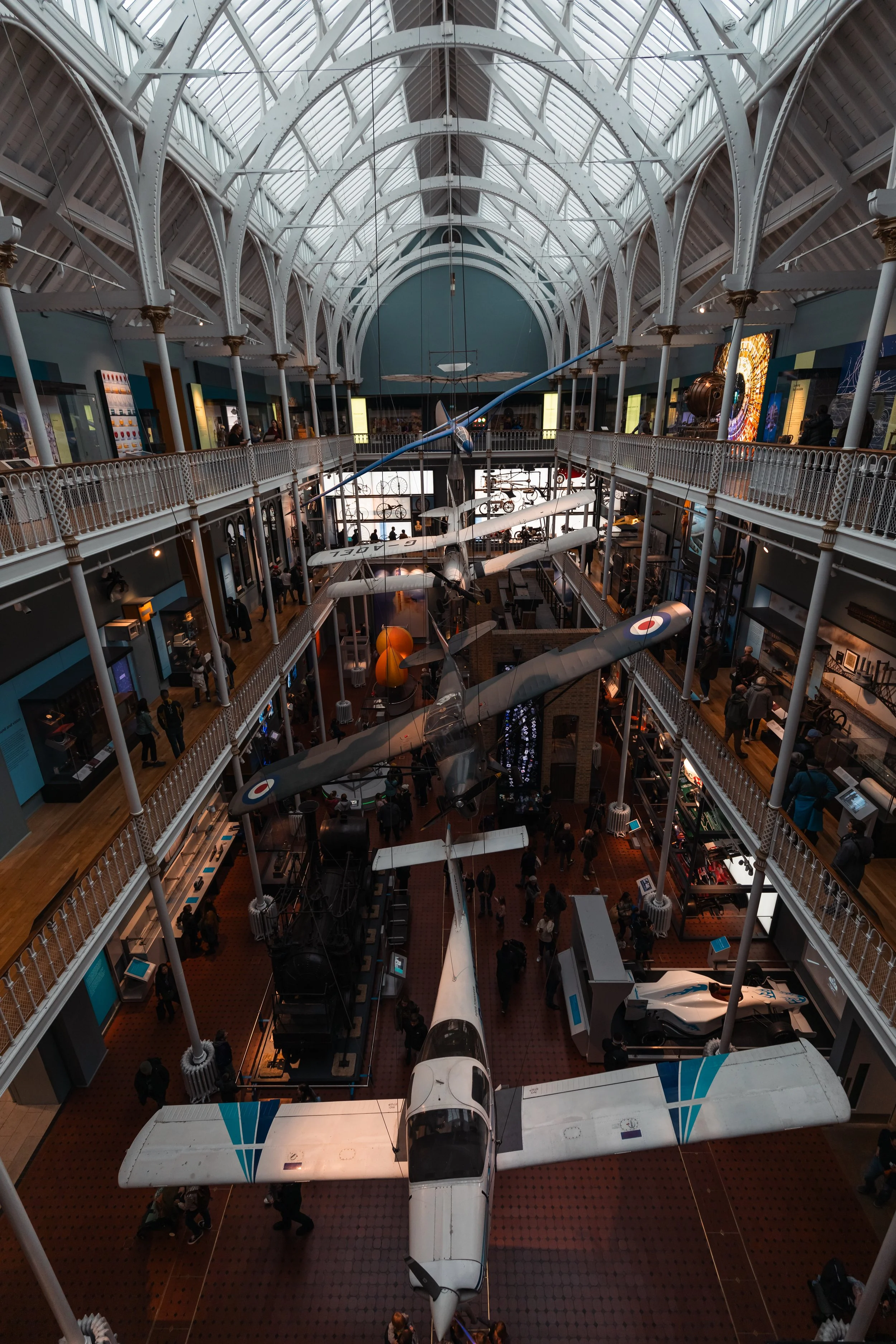 The image shows the interior of a museum or exhibit space with multiple floors. There are vintage aircraft hanging from the ceiling and various displays on the walls and floor, with visitors walking around and observing.