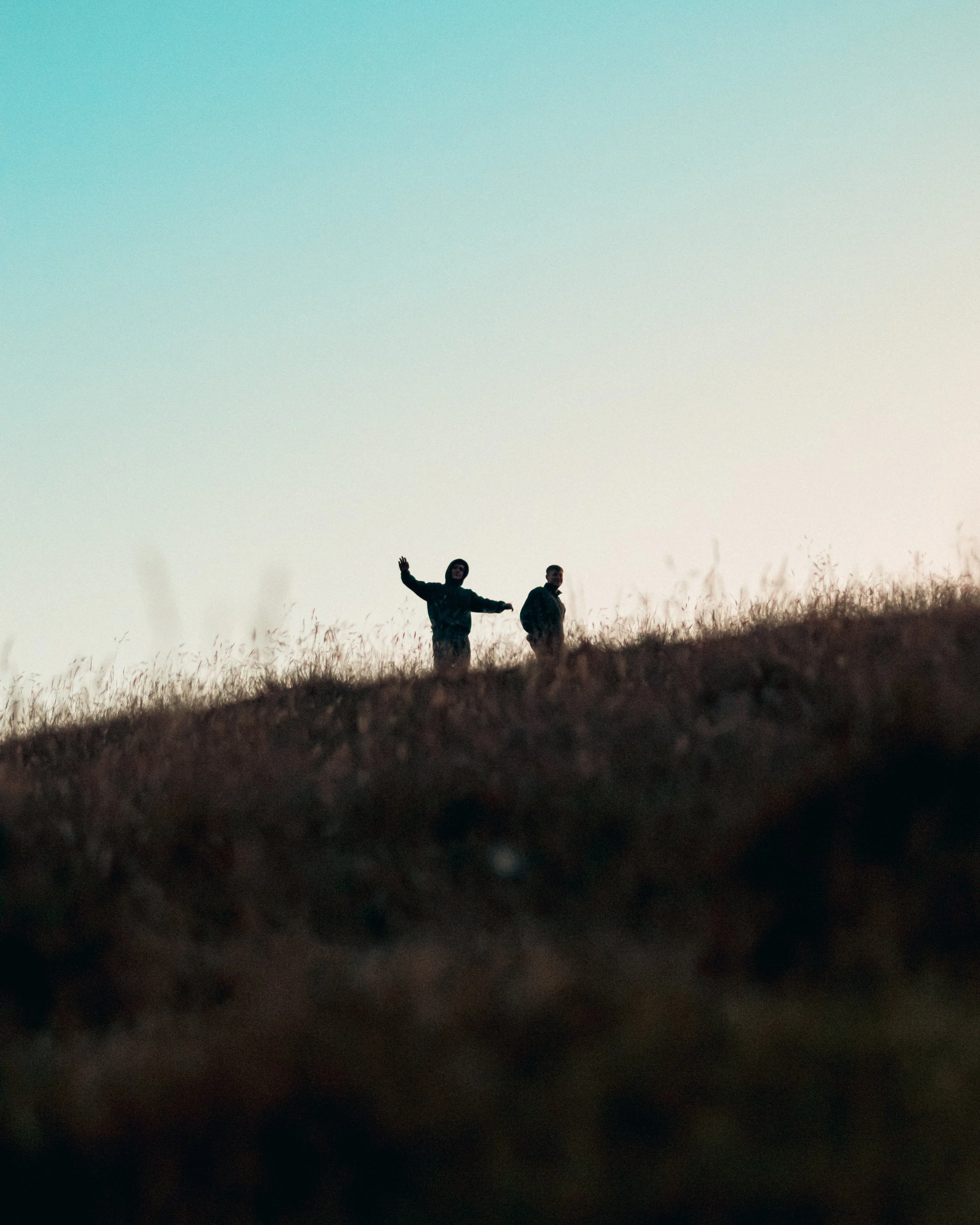 People standing on a hilltop at sunset, waving, with grass in the foreground and a colorful sky in the background.