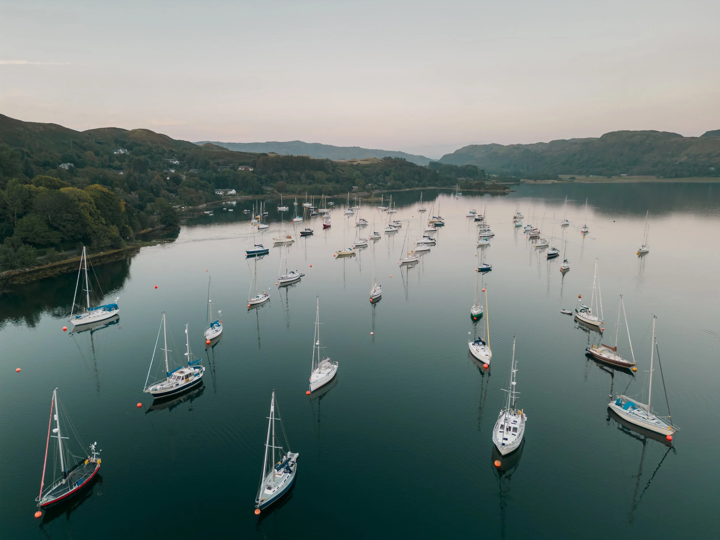 A serene harbor scene with multiple sailboats docked on calm water, surrounded by lush green hills and mountains under a cloudy sky.