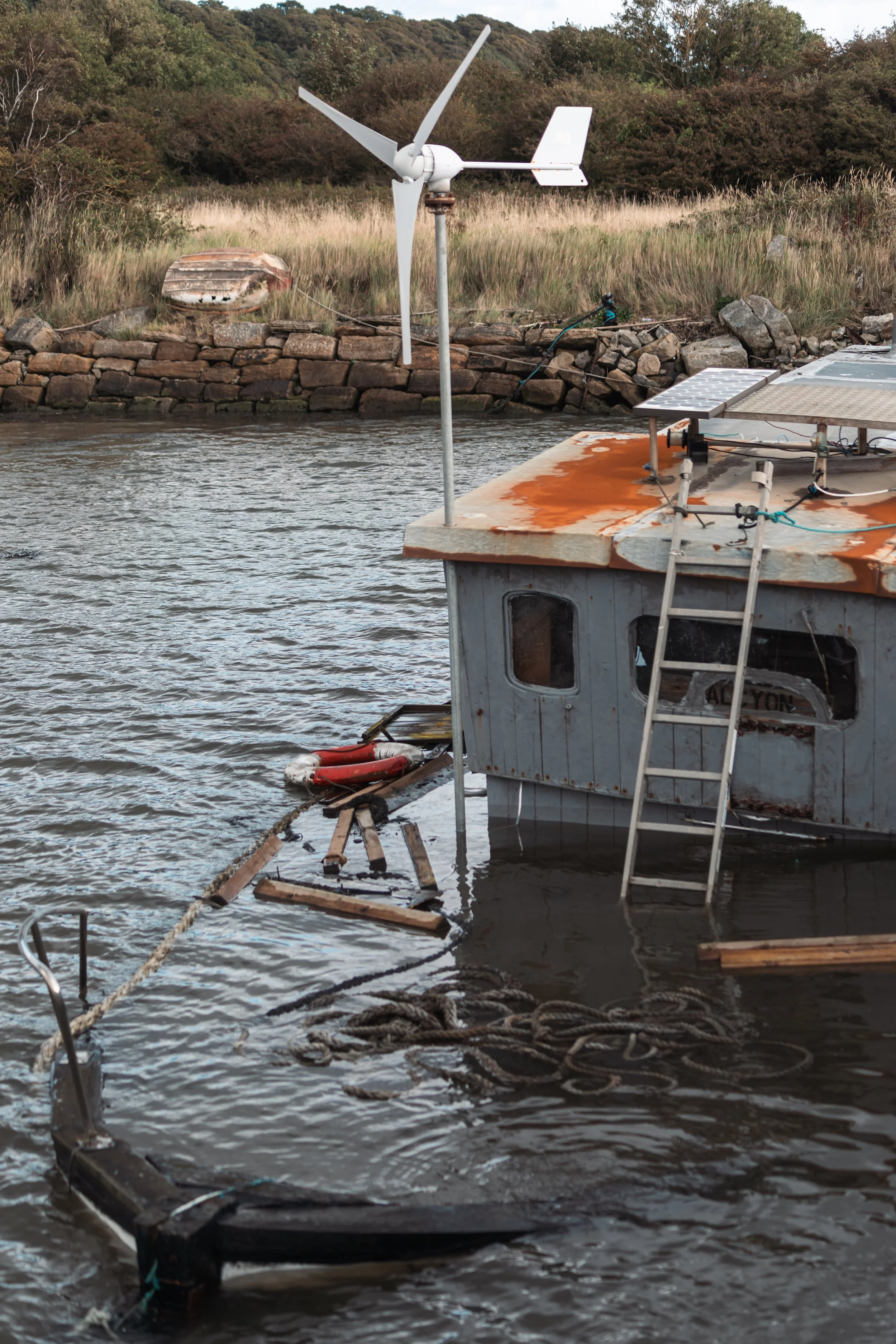 A boat on water with solar panels on the roof, a small wind turbine, a life ring, and some wooden debris floating nearby. There is a stone wall, grass, shrubs, and trees in the background.