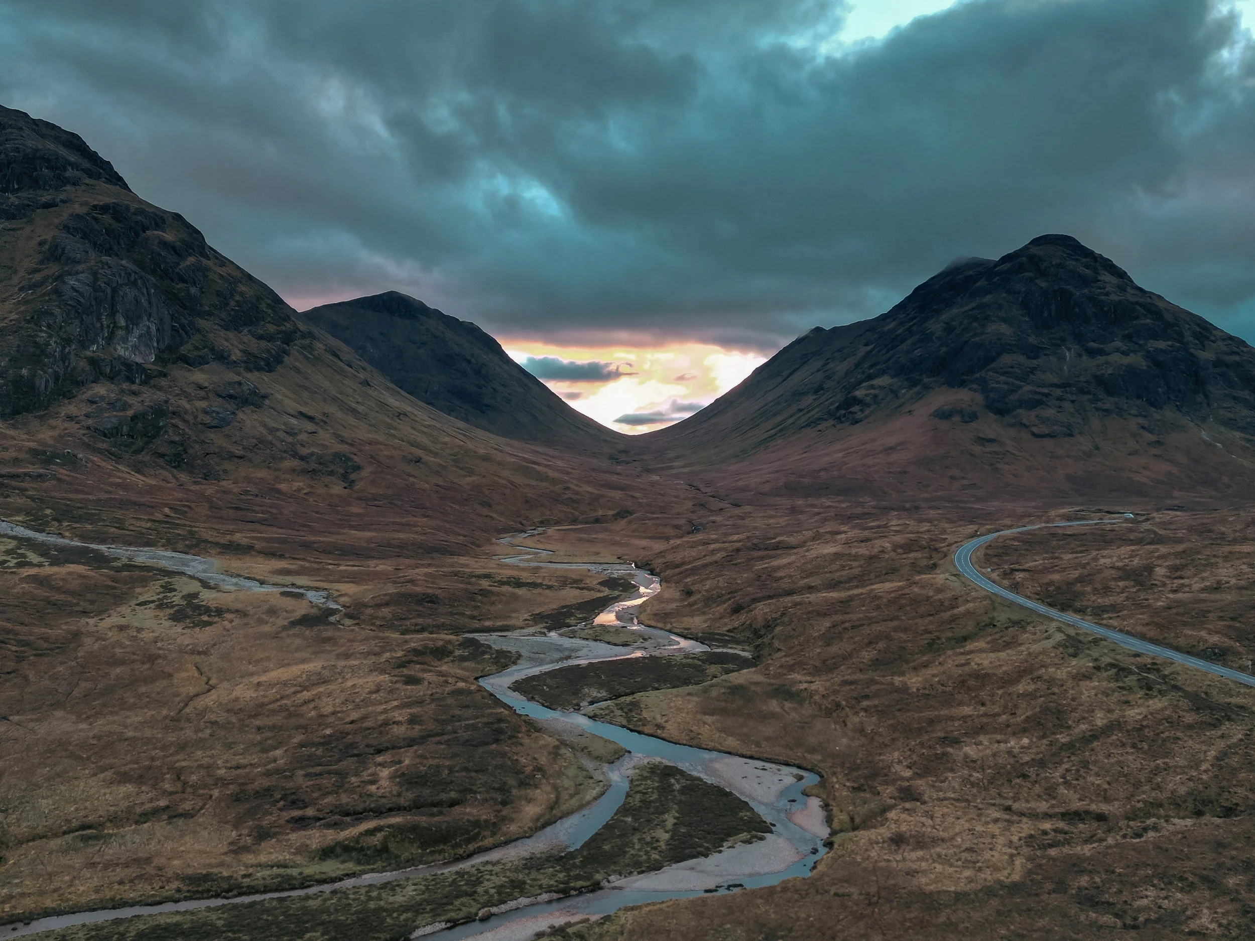 A mountainous landscape at sunset with dark clouds overhead, a winding river in the valley, and a mountain road visible in the lower right corner.