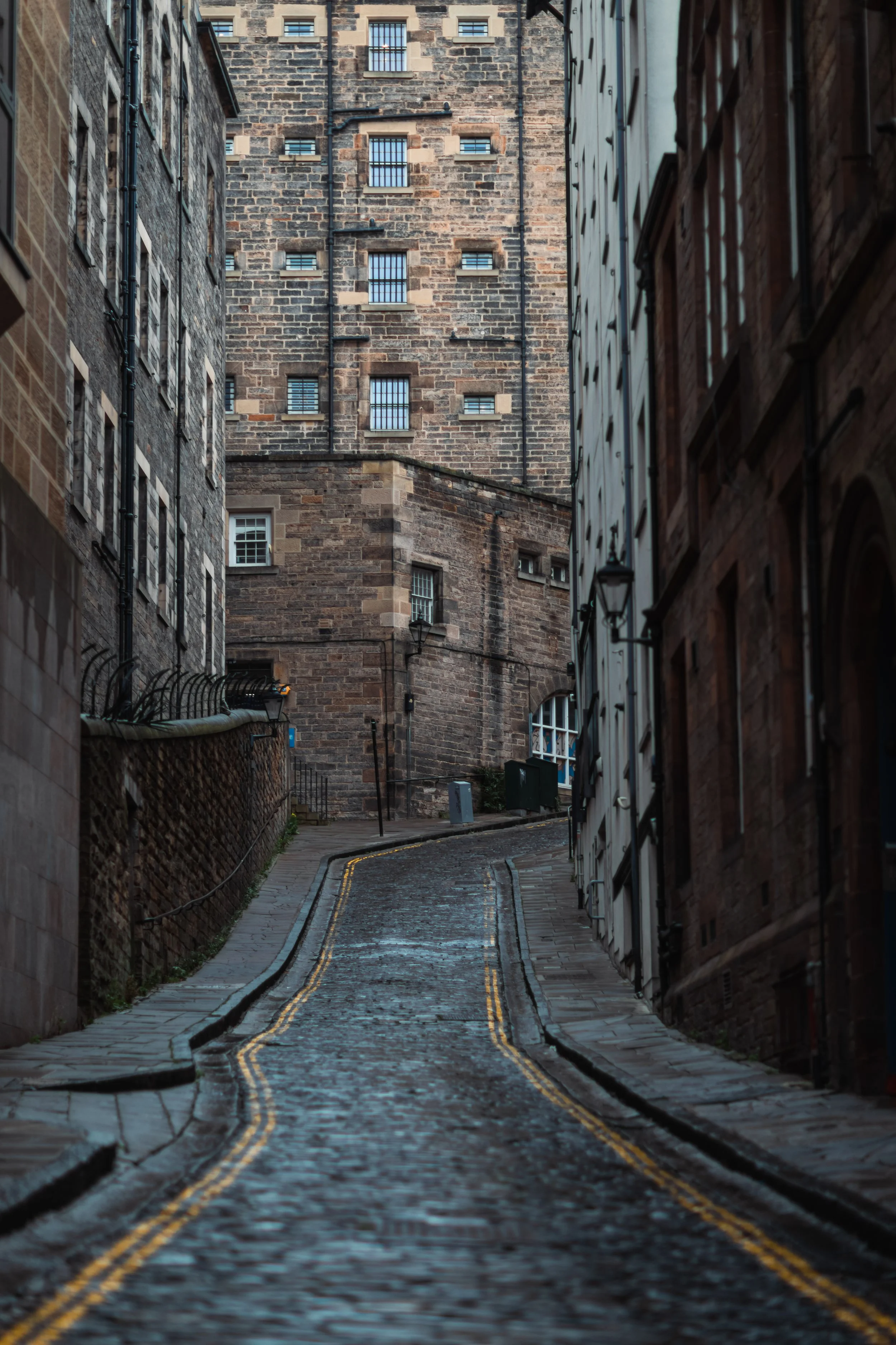 A narrow cobblestone street in an old city, lined with brick buildings on both sides, with some windows barred and street lamps mounted on the walls, leading uphill and curving out of view.