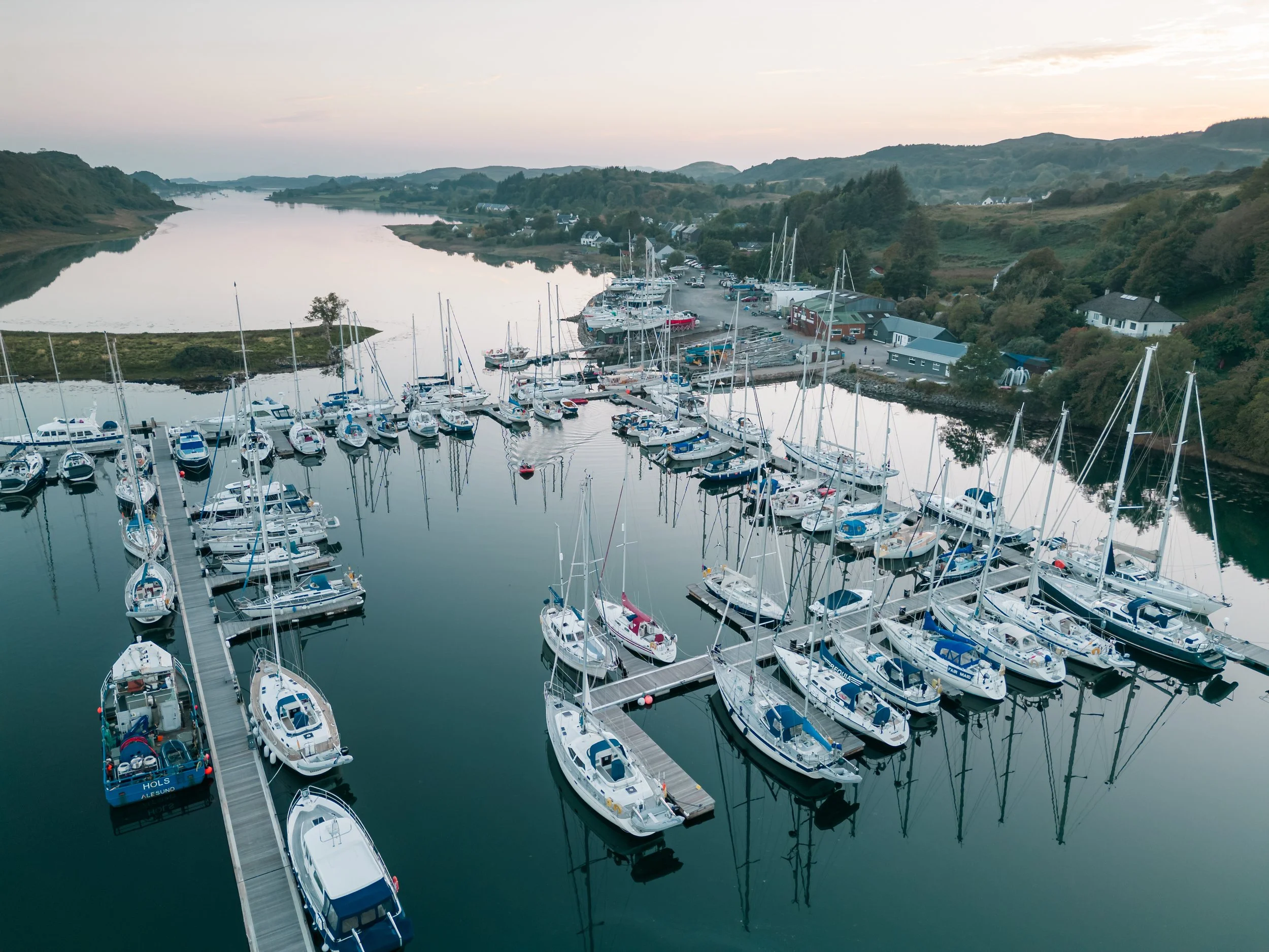 A marina with numerous sailboats and yachts docked along the piers, set in a scenic landscape with lush green hills and a calm river under an overcast sky.