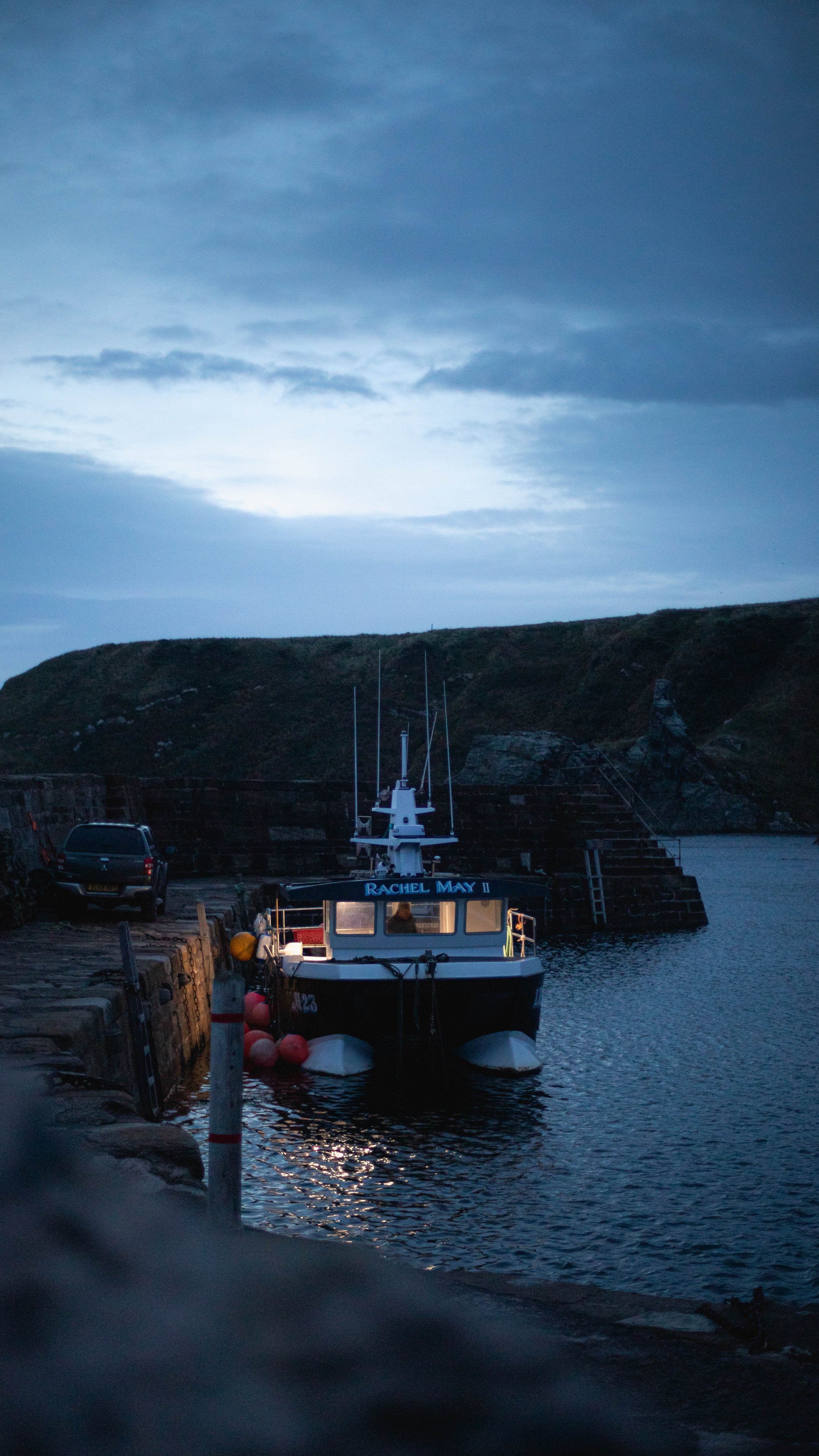 A small boat named Rachel May II docked at a pier with a person on board, under a cloudy sky during dusk or dawn, with hilly land in the background.