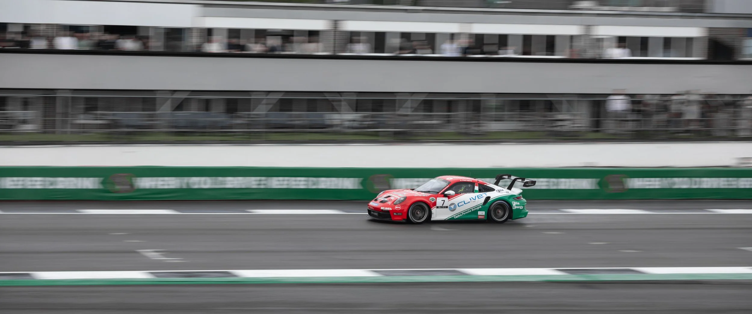 A red and white race car with the number 7 speeding on a race track, with spectators and indoor seating in the background.