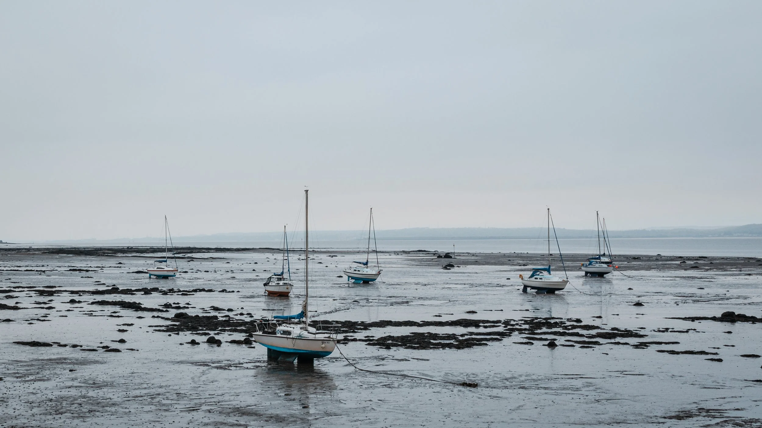 Several sailboats on a wet, rocky shoreline during low tide under a cloudy sky.