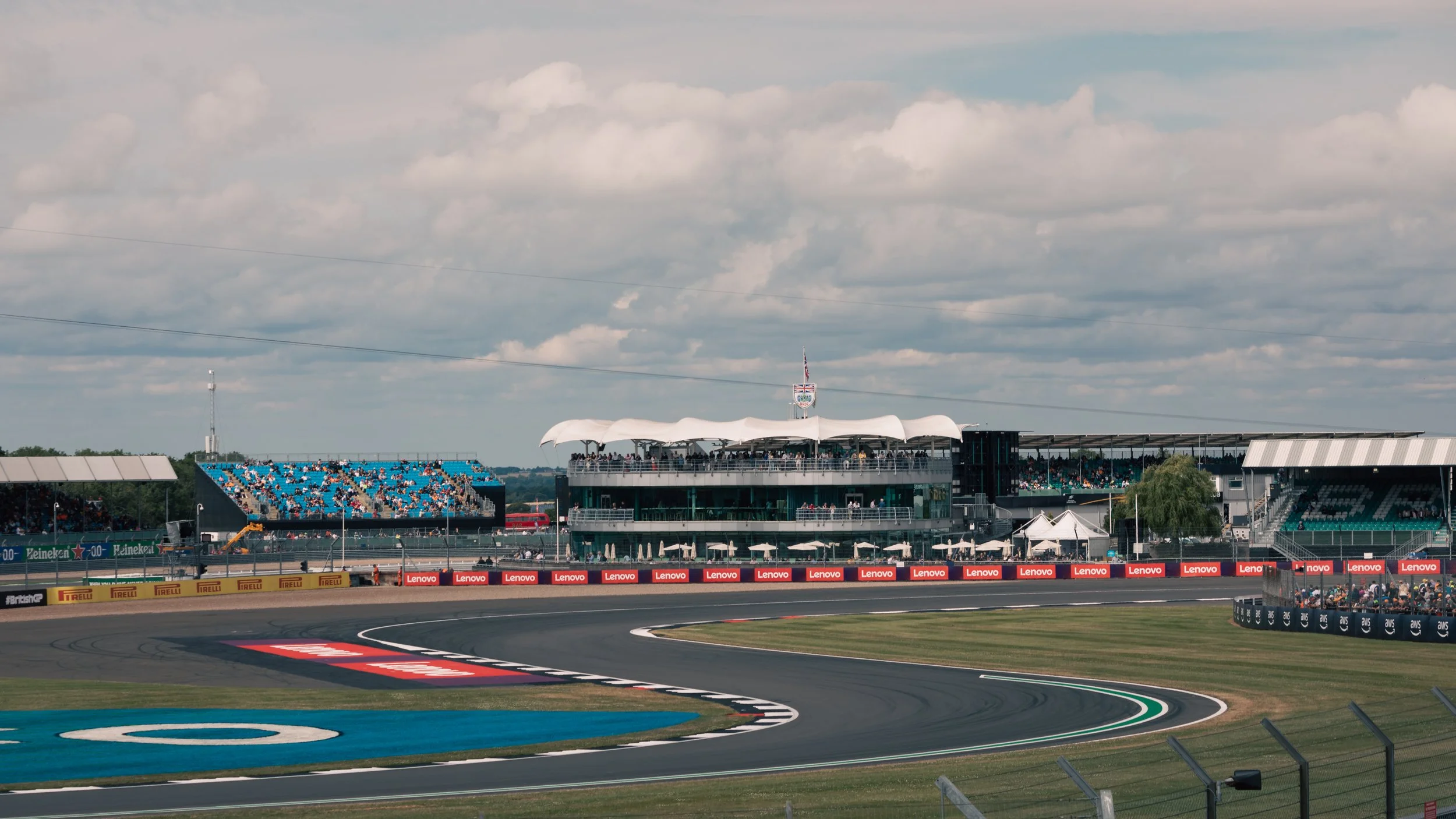 A view of a racing circuit with stands filled with spectators, a race track with painted lines, and a modern grandstand with a white canopy roof, under a cloudy sky.