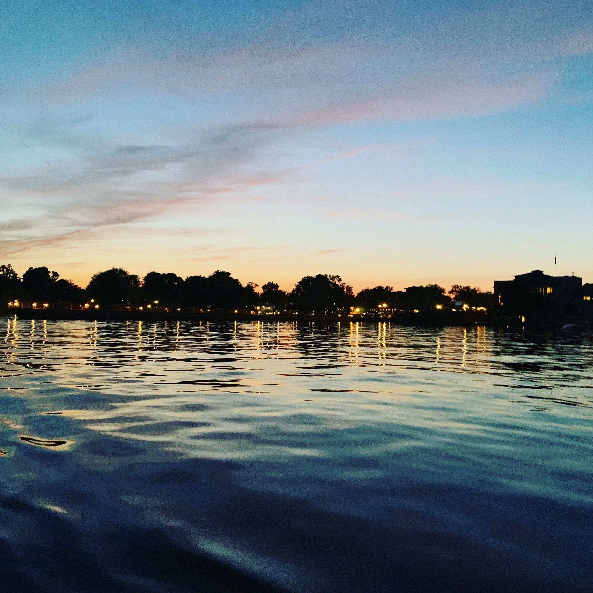 Skaneateles Lake at sunset reflecting the colors of a sunset with trees and buildings along the shoreline and a pastel-colored sky with clouds.