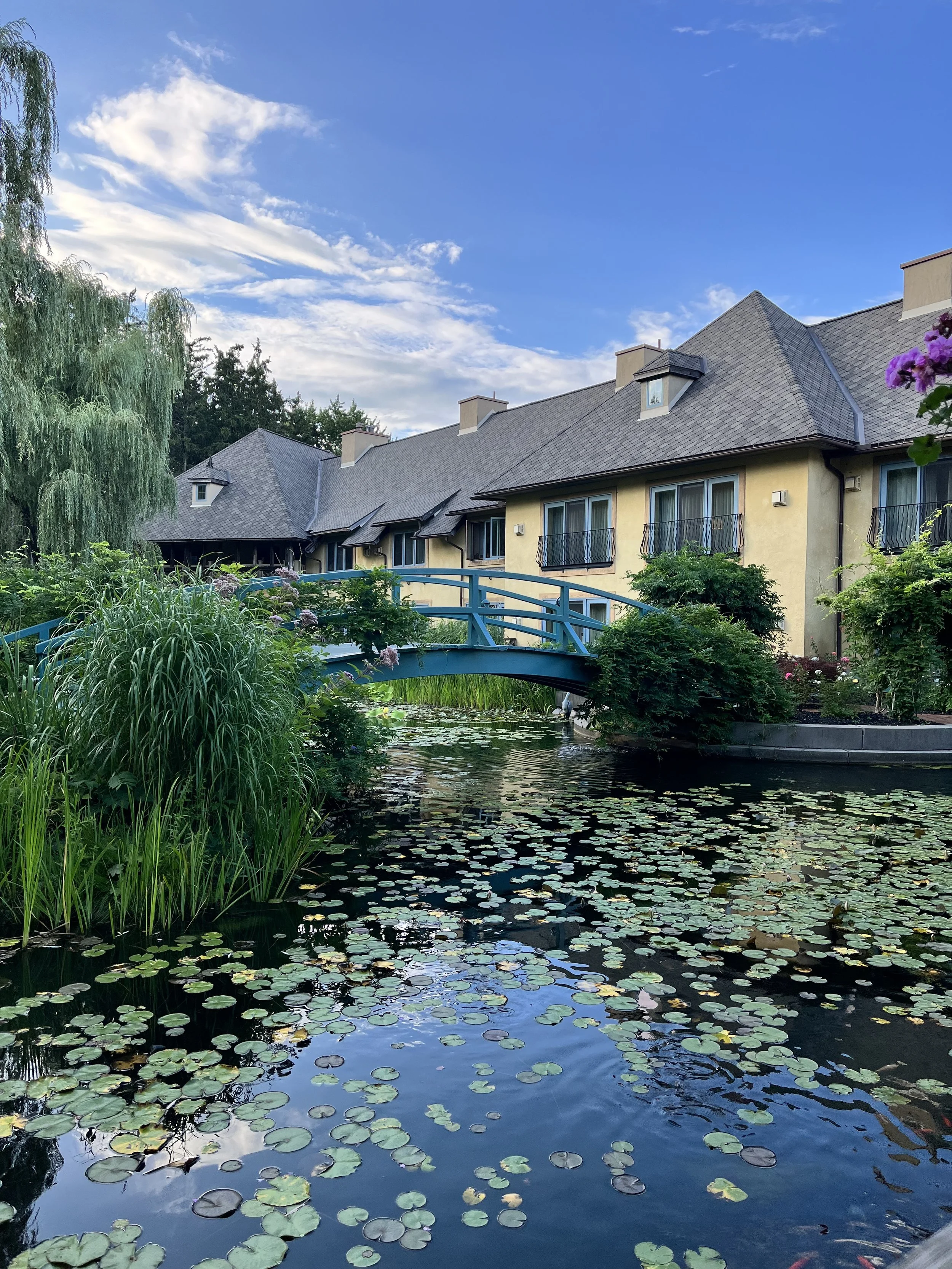 A serene outdoor scene with a small pond covered in water lilies in front of a yellow residential building with multiple windows and balconies. This is Mirbeau Inn and Spa in Skaneateles, NY. It is inspired by Monet's gardens.