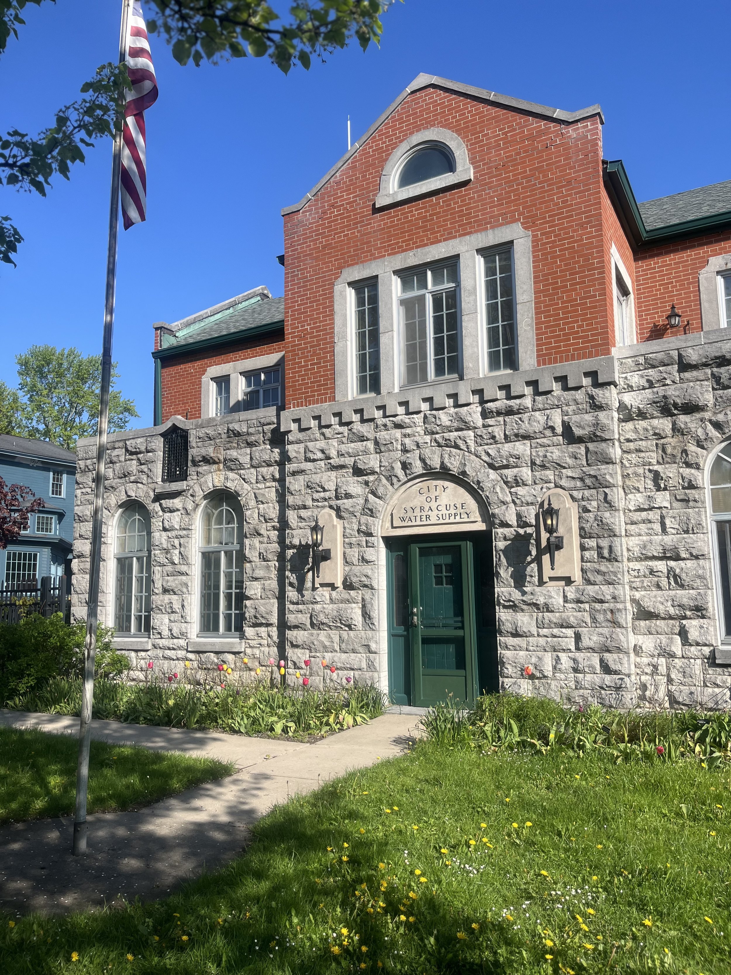 A stone and brick building with a green door, arched windows, and a sign that reads "City of Syracuse Water Supply." There is a sidewalk leading to the entrance, a grassy area with tulips and other flowers, and an American flag on a pole in front of the building.