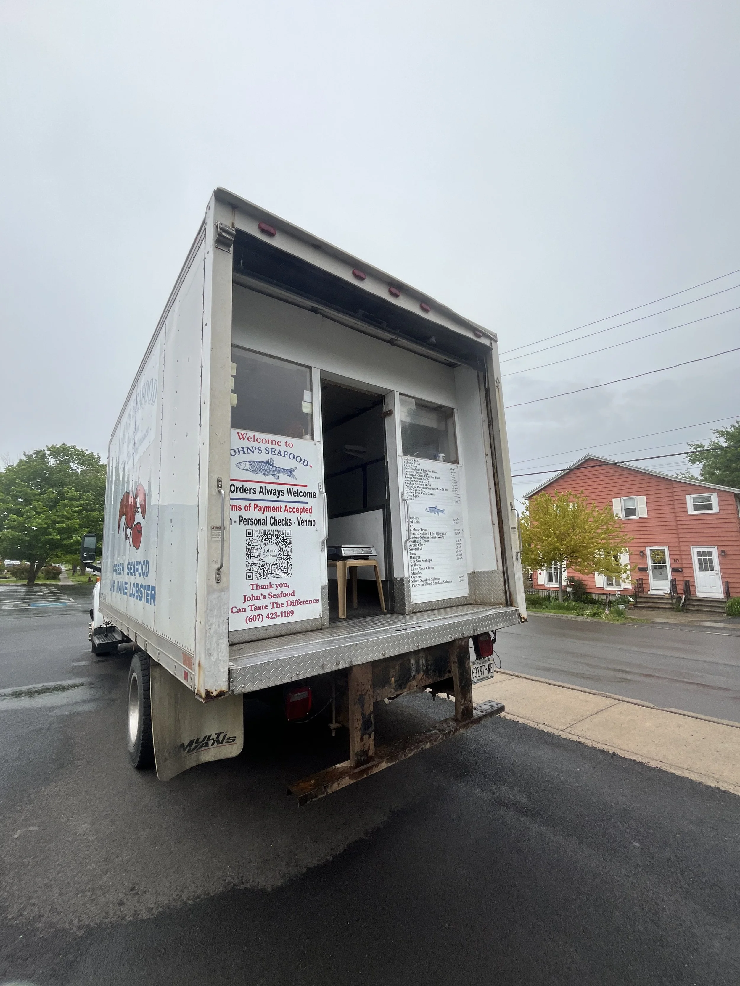 Food truck with open service window, signboard displaying menu, and two small stools outside, parked on street with houses and trees in the background in the village of Skaneateles, New York.