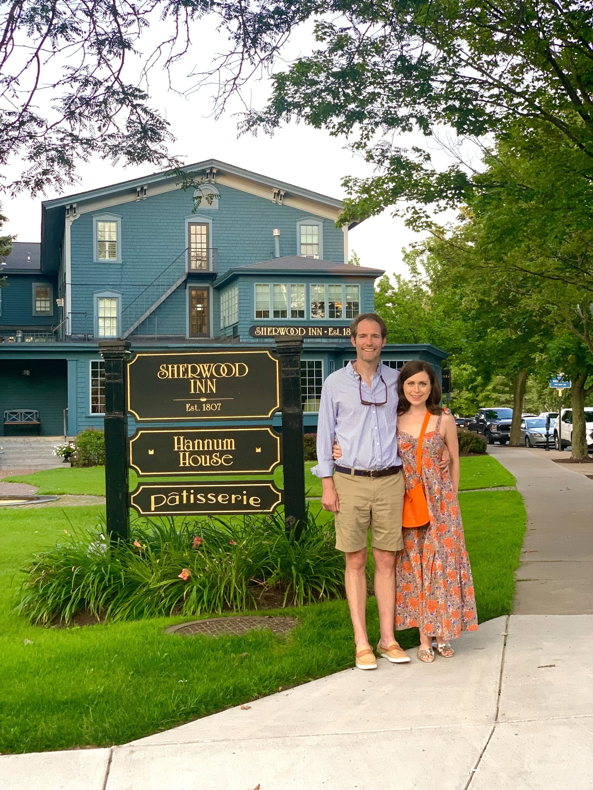 Two people standing in front of the Sherwood Inn sign in Skaneateles, New York with a blue building and trees in the background. The man is wearing a light purple striped shirt and shorts, the woman is wearing a colorful floral dress and sandals.