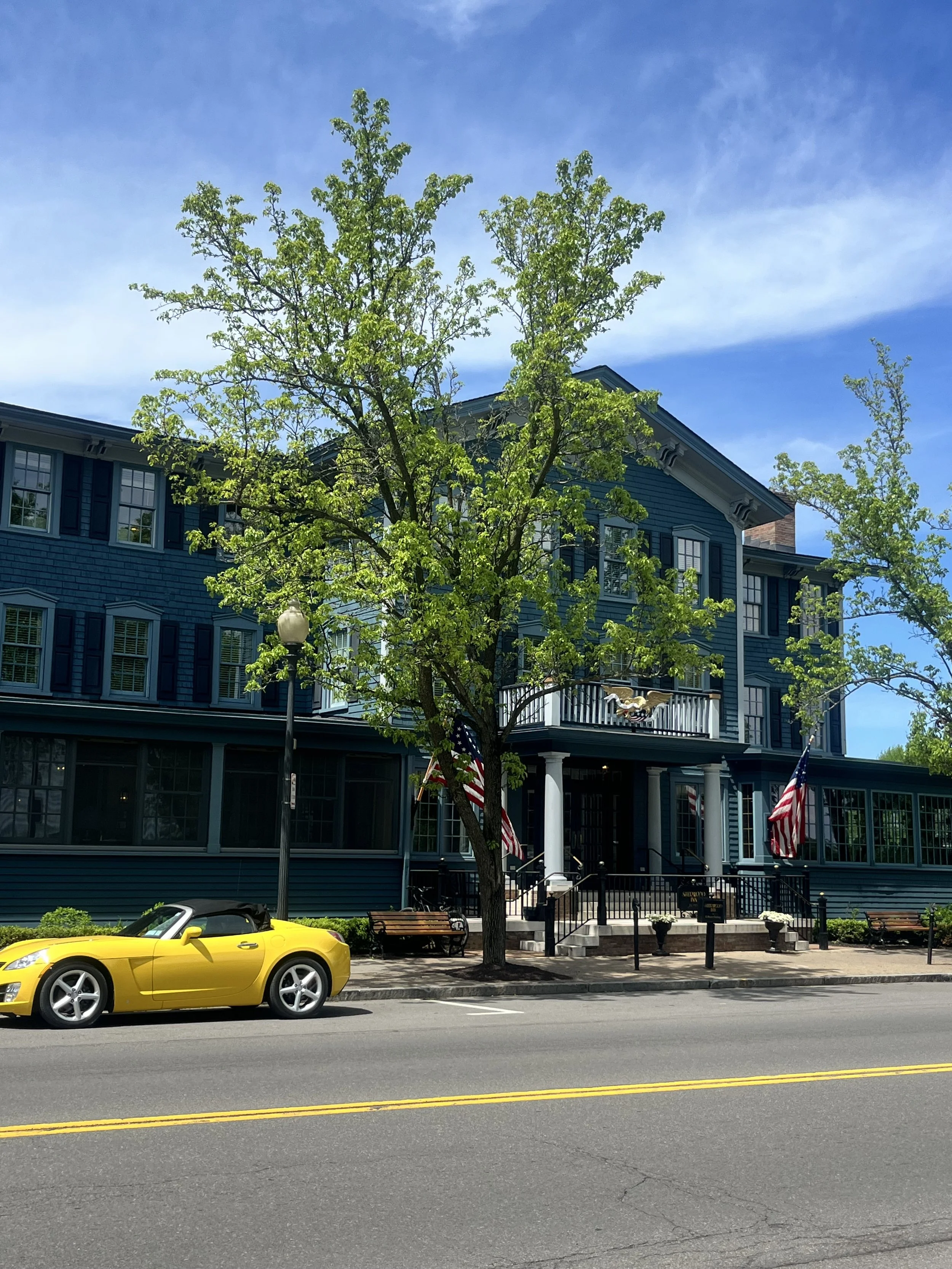 Yellow sports car parked on the street in front of a blue house with a porch, multiple windows, and American flags, with a tree and a lamp post in the scene, under a partly cloudy sky. This is the Sherwood Inn in Skaneateles, NY.