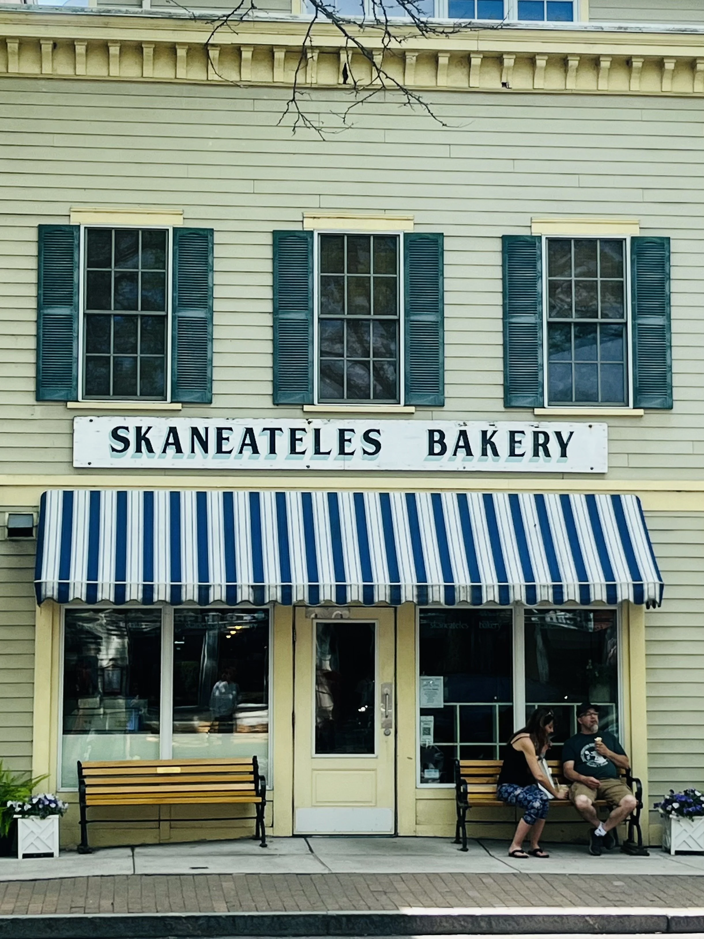 Storefront of Skaneateles Bakery with yellow exterior, blue shutters on upper windows, striped blue and white awning, and two benches outside. Two people are sitting on the bench near the entrance, conversing with each other.