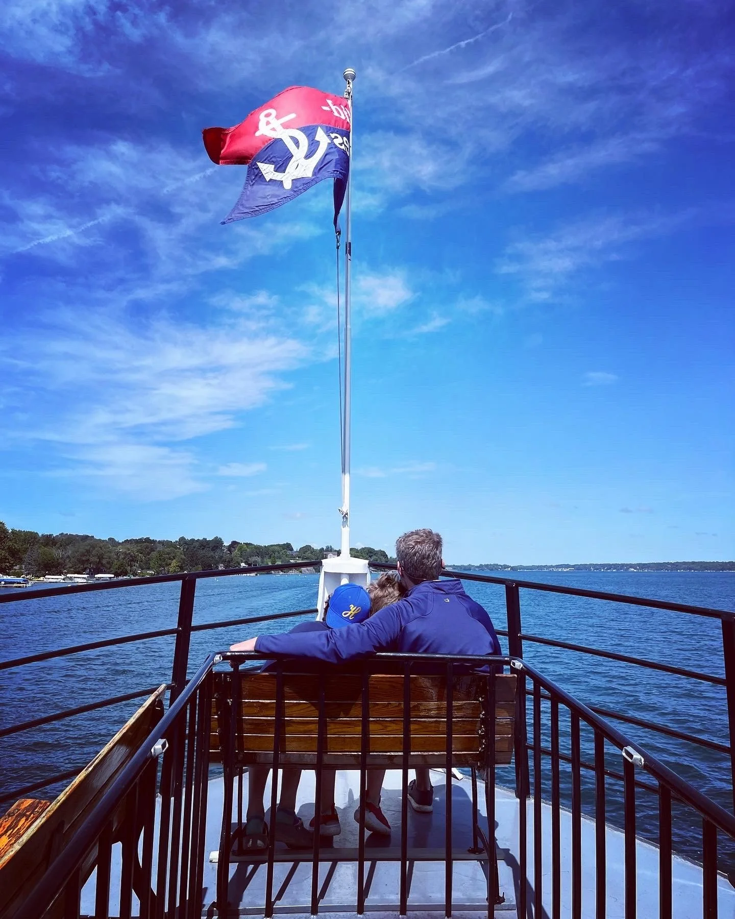People sitting on a boat enjoying a scenic water view under a blue sky with wispy clouds, a flag flying from the boat's mast in the background. They are on the Judge Ben boat in Skaneateles, NY.