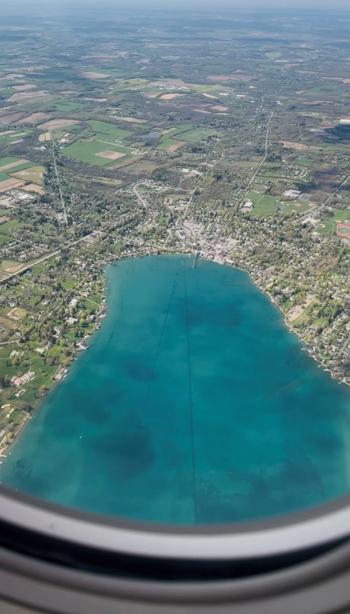 Aerial view of Skaneateles Lake from an airplane window, showing residential neighborhoods, a lake, and surrounding farmland.