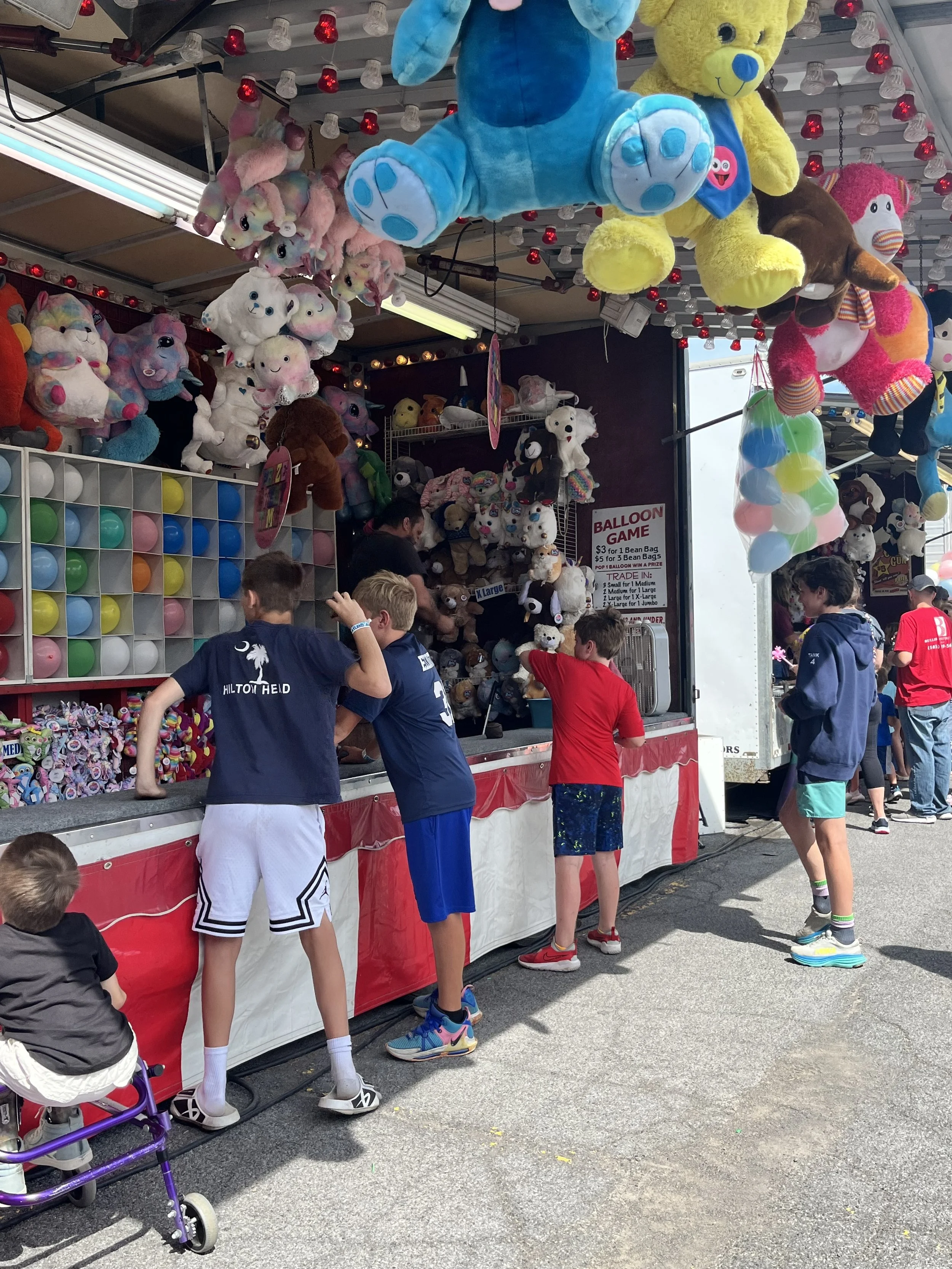 Children playing a balloon game at a fair or carnival, with various large plush toys hanging overhead. The game involves popping balloons, and an adult is assisting the children. They are at the Skaneateles Field Days in New York.