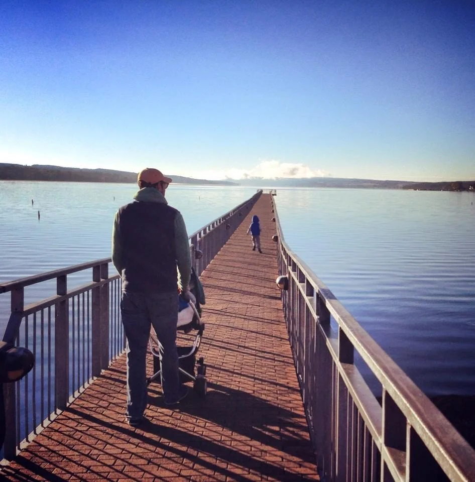 A man pushes a stroller on a long pier out over calm water, with a child walking ahead towards the horizon, under a clear blue sky in Skaneateles, NY on the pier.