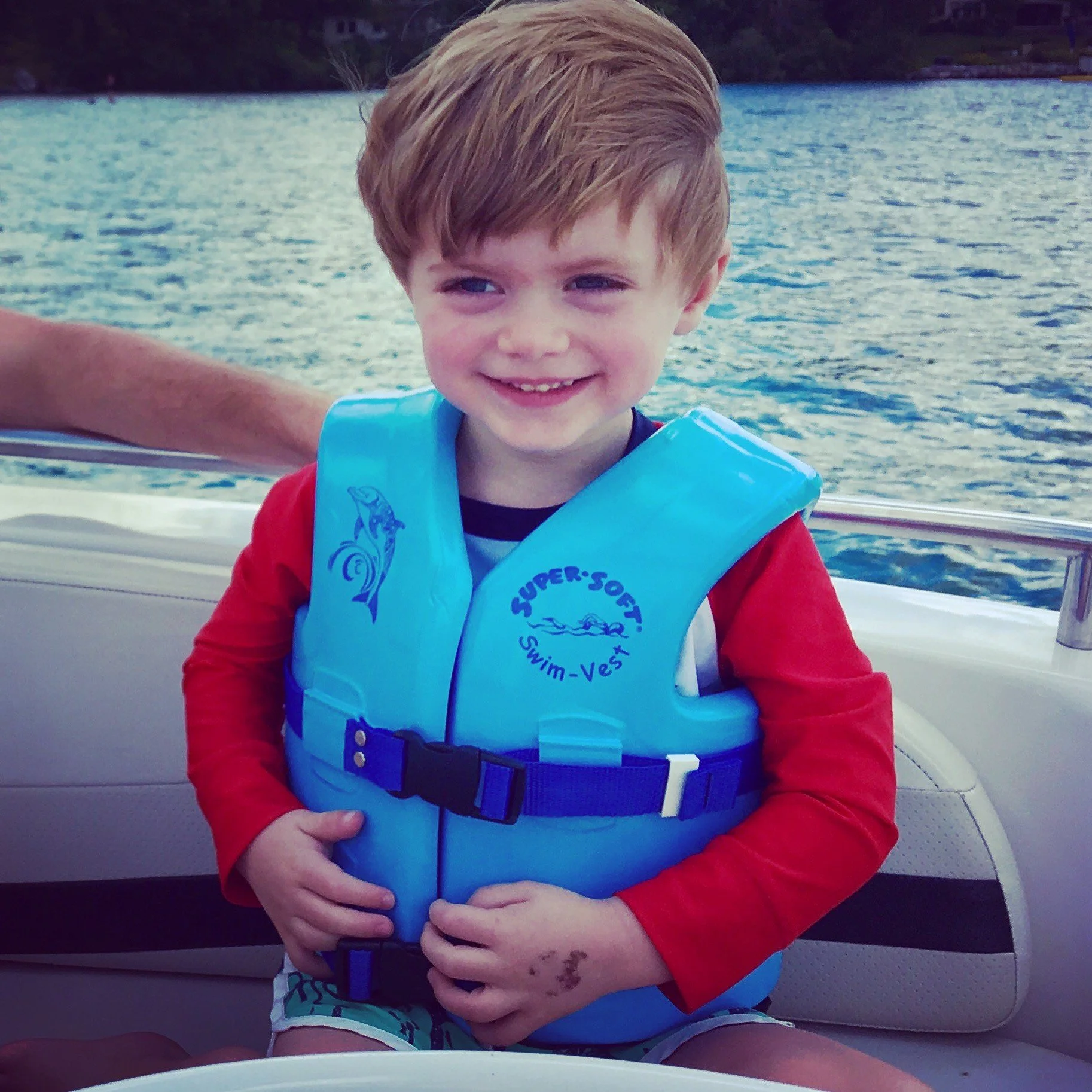 A young boy sitting on a boat in Skaneateles wearing a blue life jacket and red shirt, smiling with a body of water and trees in the background.