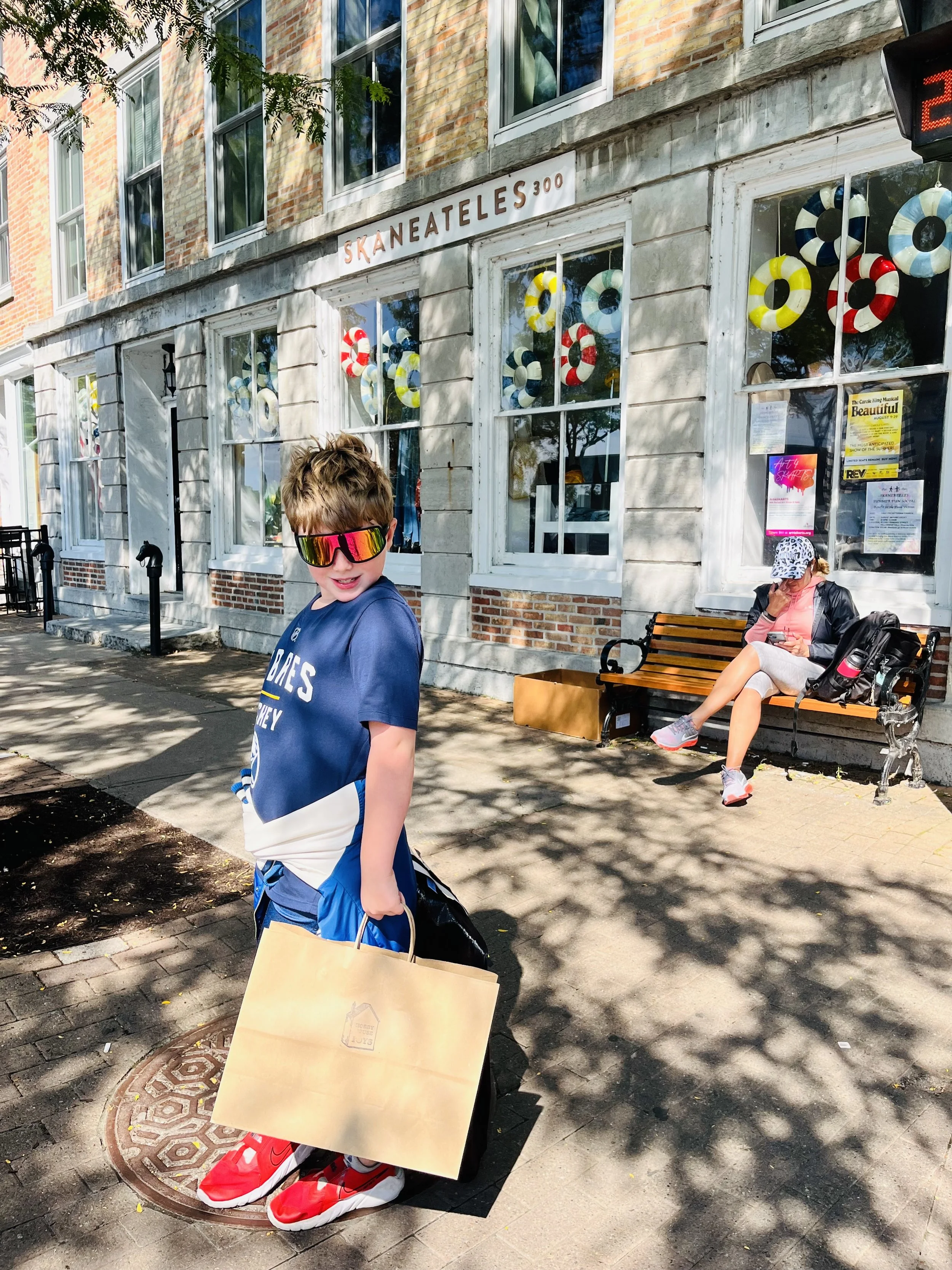 A young boy wearing red sneakers, sunglasses, a blue and white sports shirt, and blue shorts holding a shopping bag standing on a sidewalk. In the background, a woman in a hat and pink shirt is sitting on a park bench, looking at her phone, with a large backpack next to her. The building behind them has many windows and a sign reading 'SKANEATELES 300'.