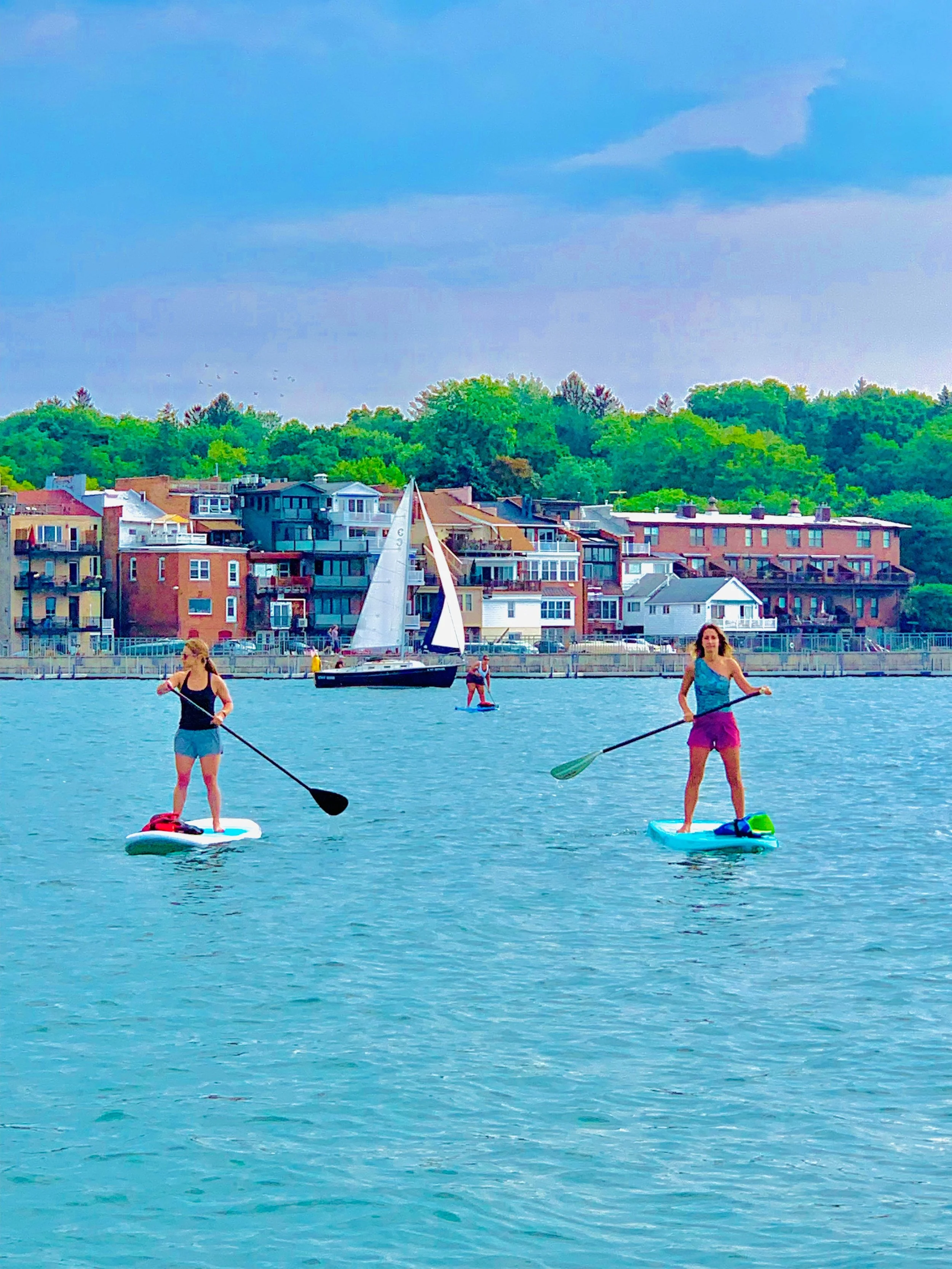 Two women paddleboarding on Skaneateles Lake with colorful buildings and green trees in the background.