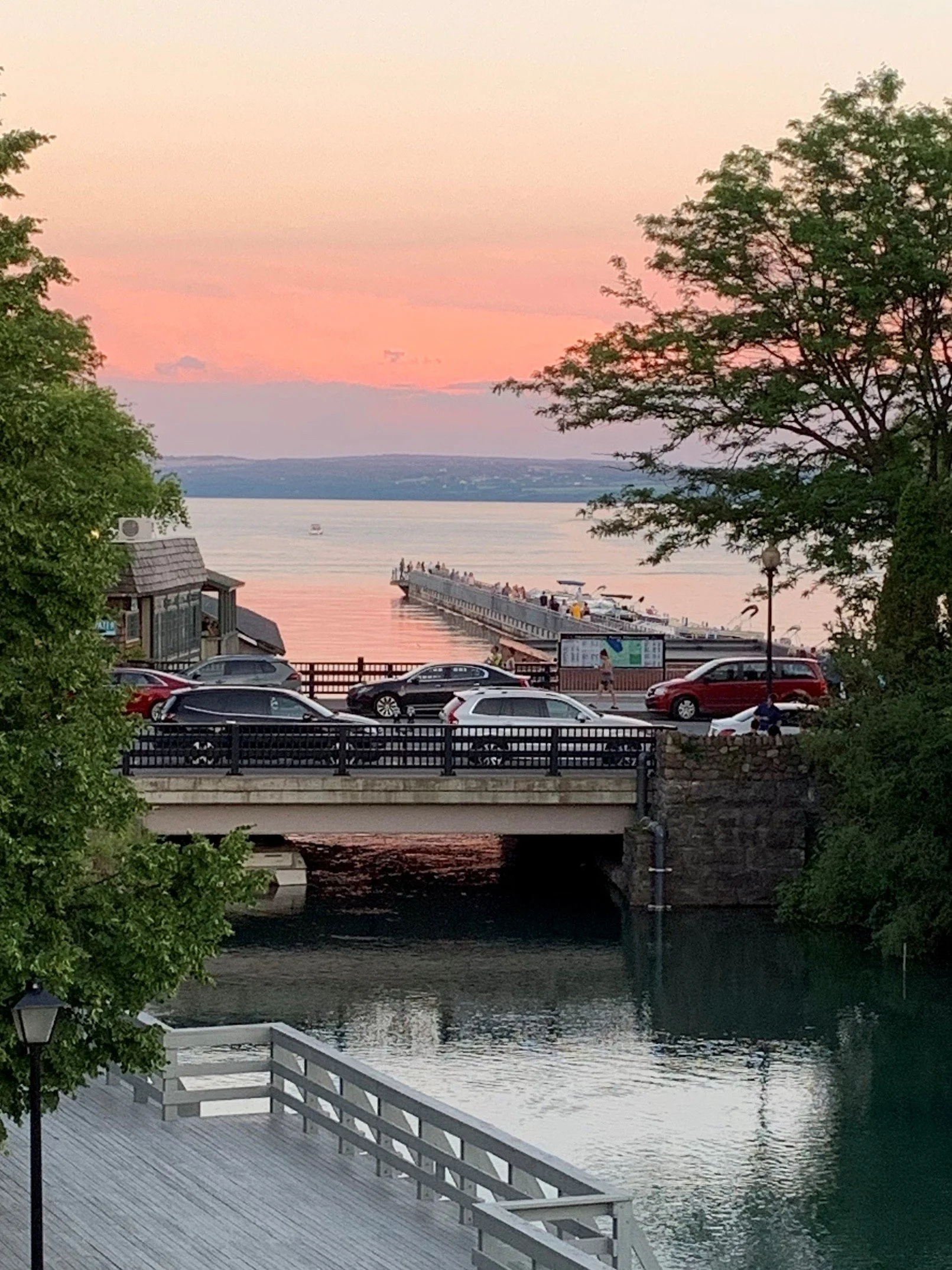 A scenic view of a waterfront in Skaneateles, New York at sunset with trees, a bridge, boats on the water, and cars parked near the water.