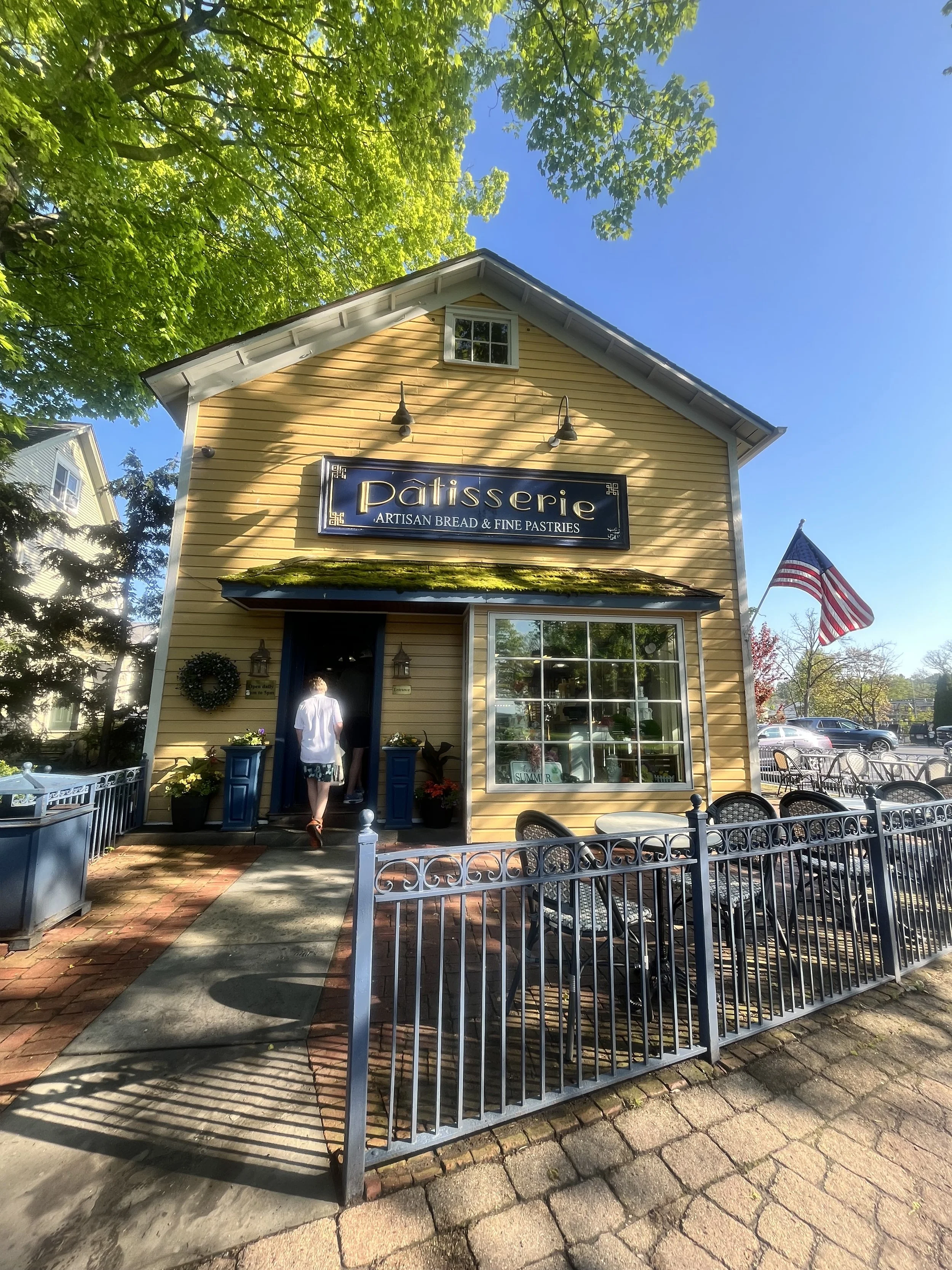 Yellow bakery storefront with sign reading 'Pâtisserie artisan bread & fine pastries' on a sunny day, American flag flying nearby, and people entering the store in Skaneateles, New York.
