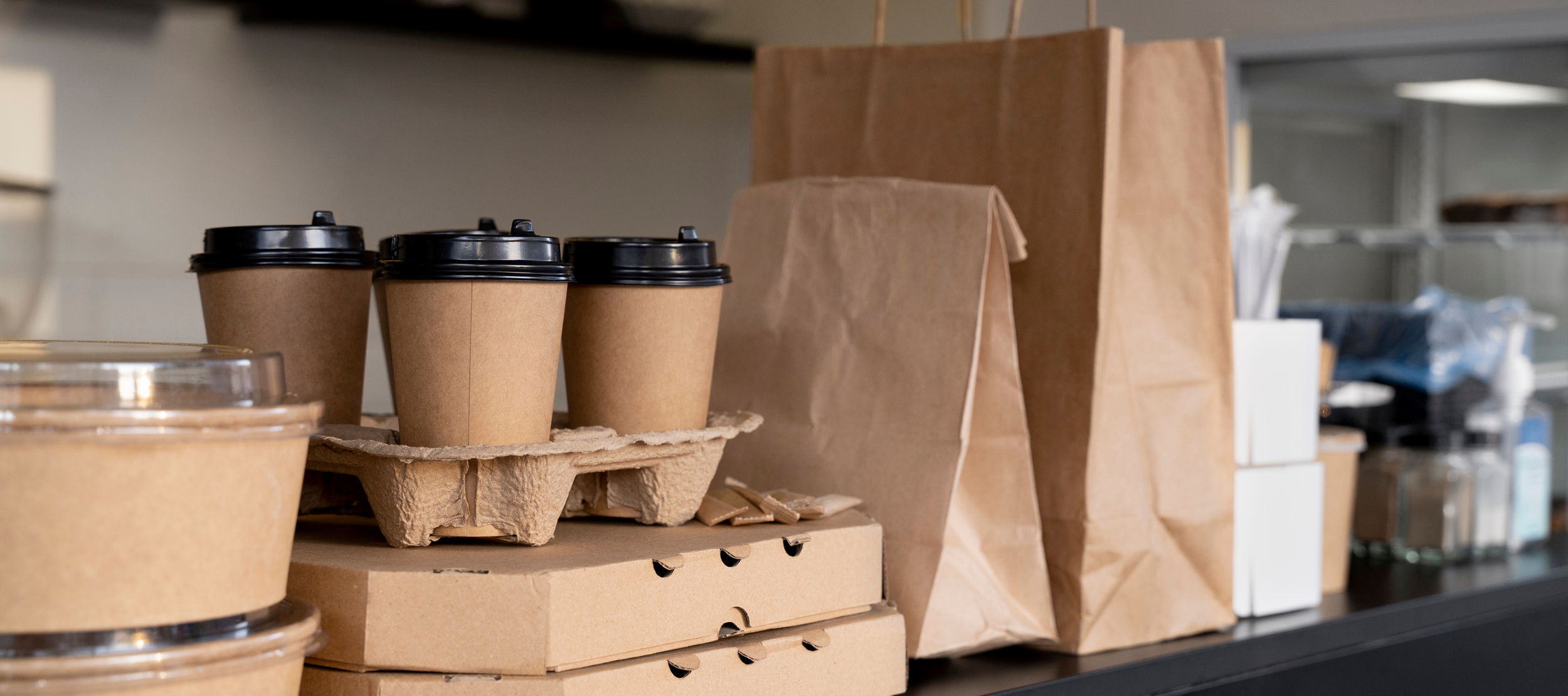 A collection of brown paper coffee cups with black lids, some stacked and some in paper bags, on a countertop in a storage room.