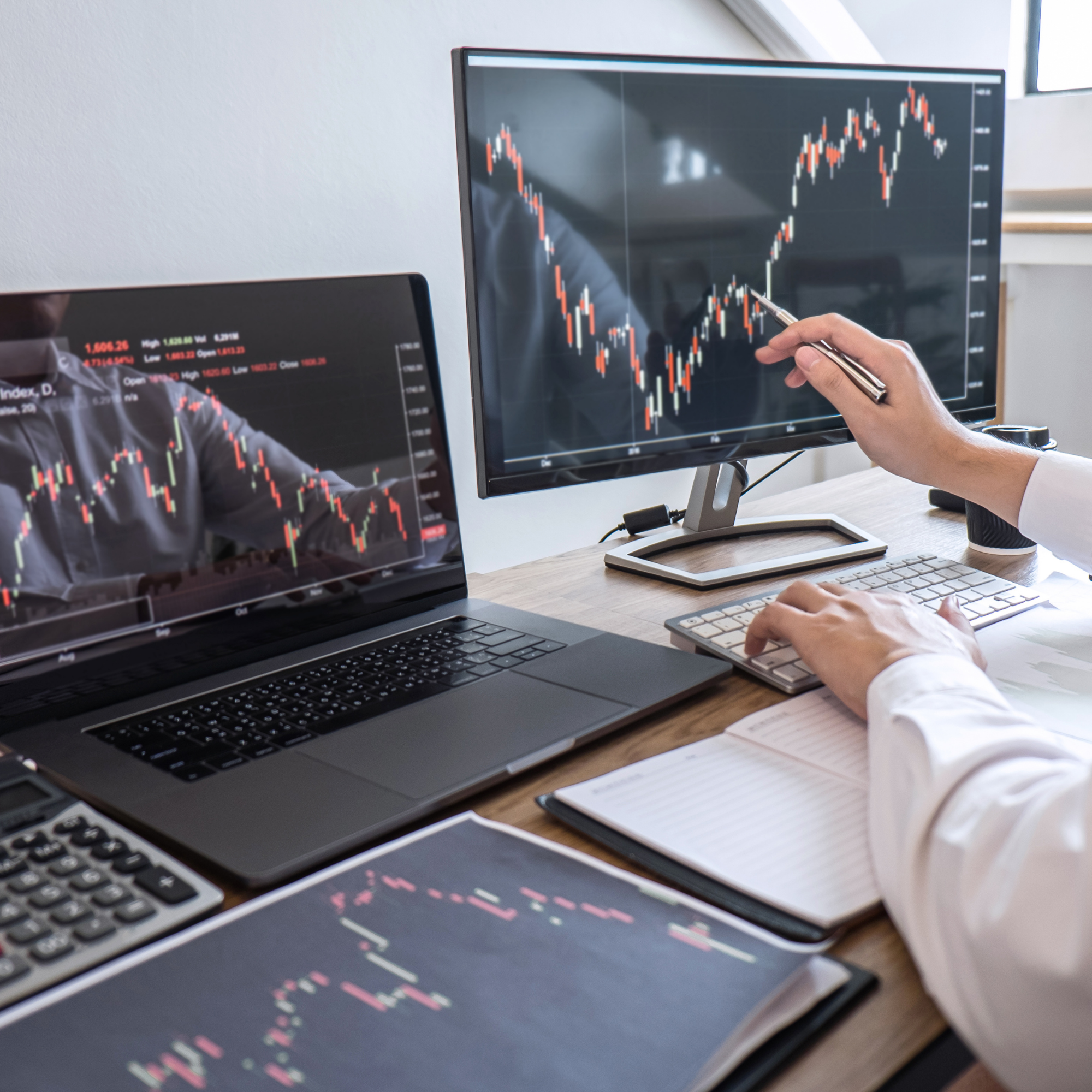 Multiple computer monitors showing stock market charts with candlestick patterns, a person's hand with a pen pointing at one of the charts, and a keyboard and papers on a wooden desk.
