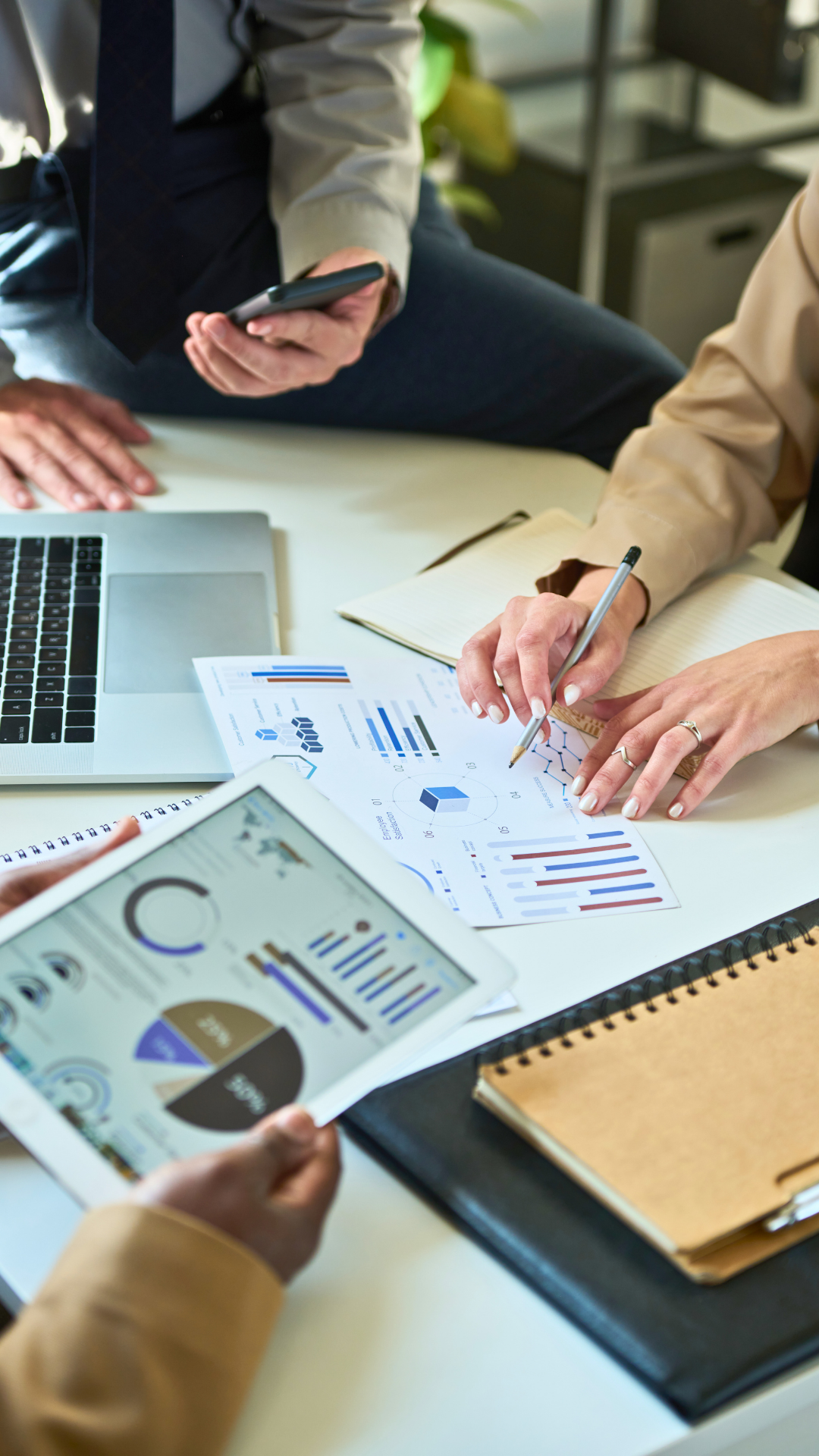 People working on business reports and analytics with charts and graphs on paper, laptop, tablet, and notebook in an office setting.