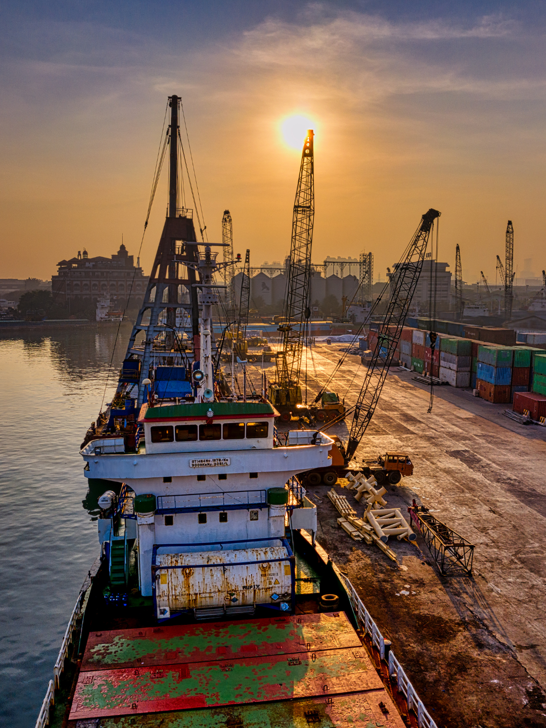 Cargo ship docked at a port during sunset, with cranes and stacked shipping containers nearby.
