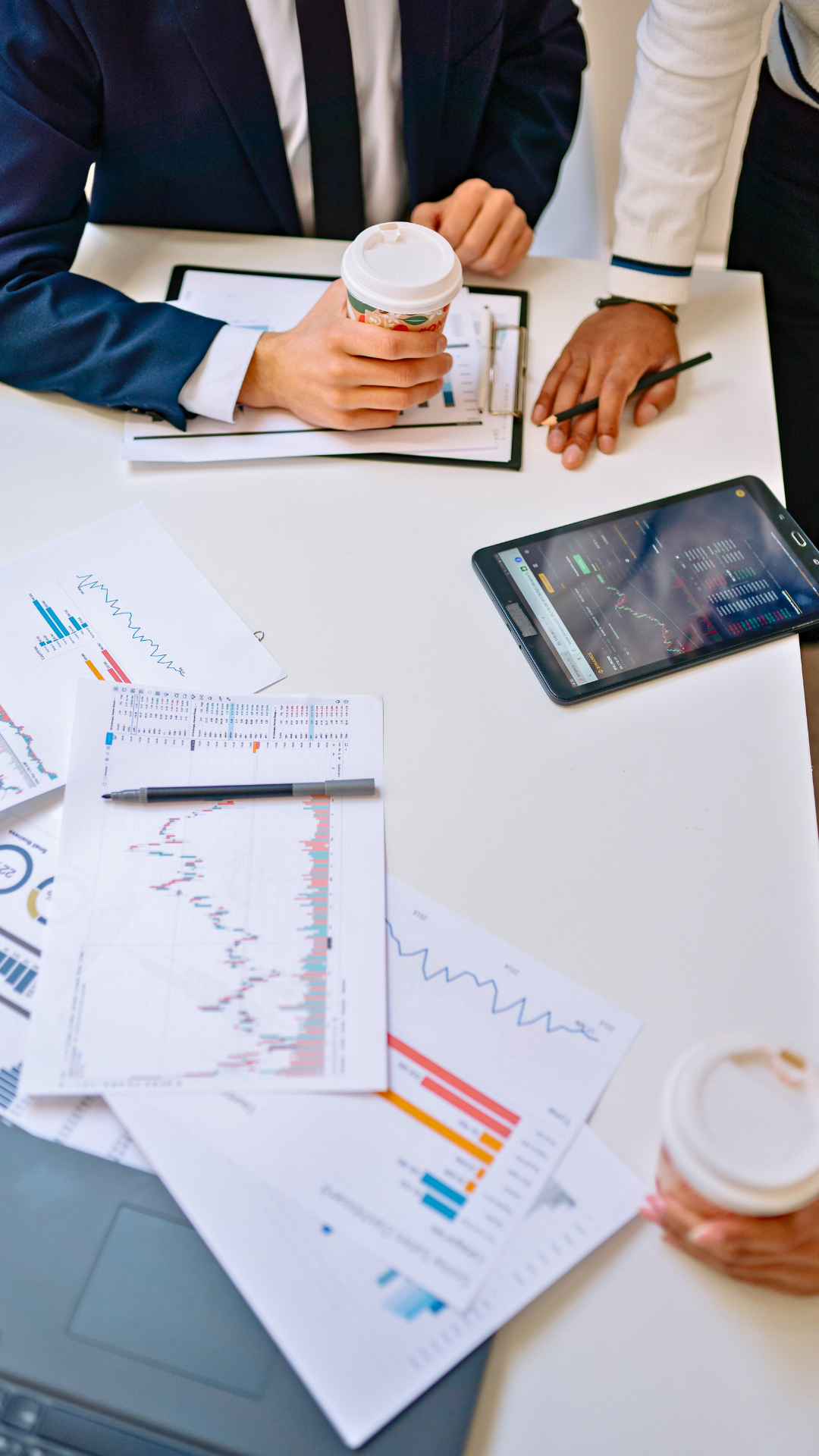 Business meeting with financial charts, graphs, a tablet displaying stock market data, and two people holding coffee cups.
