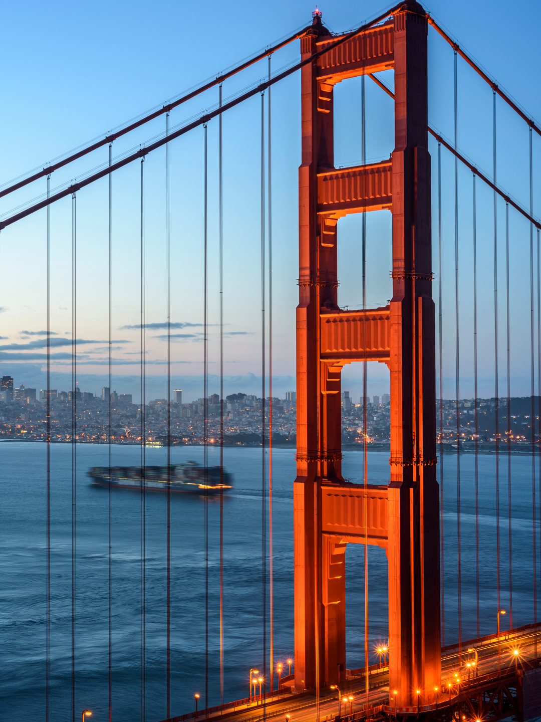 Nighttime view of the Golden Gate Bridge in San Francisco, illuminated with orange lights, with a boat passing underneath and city lights in the background.
