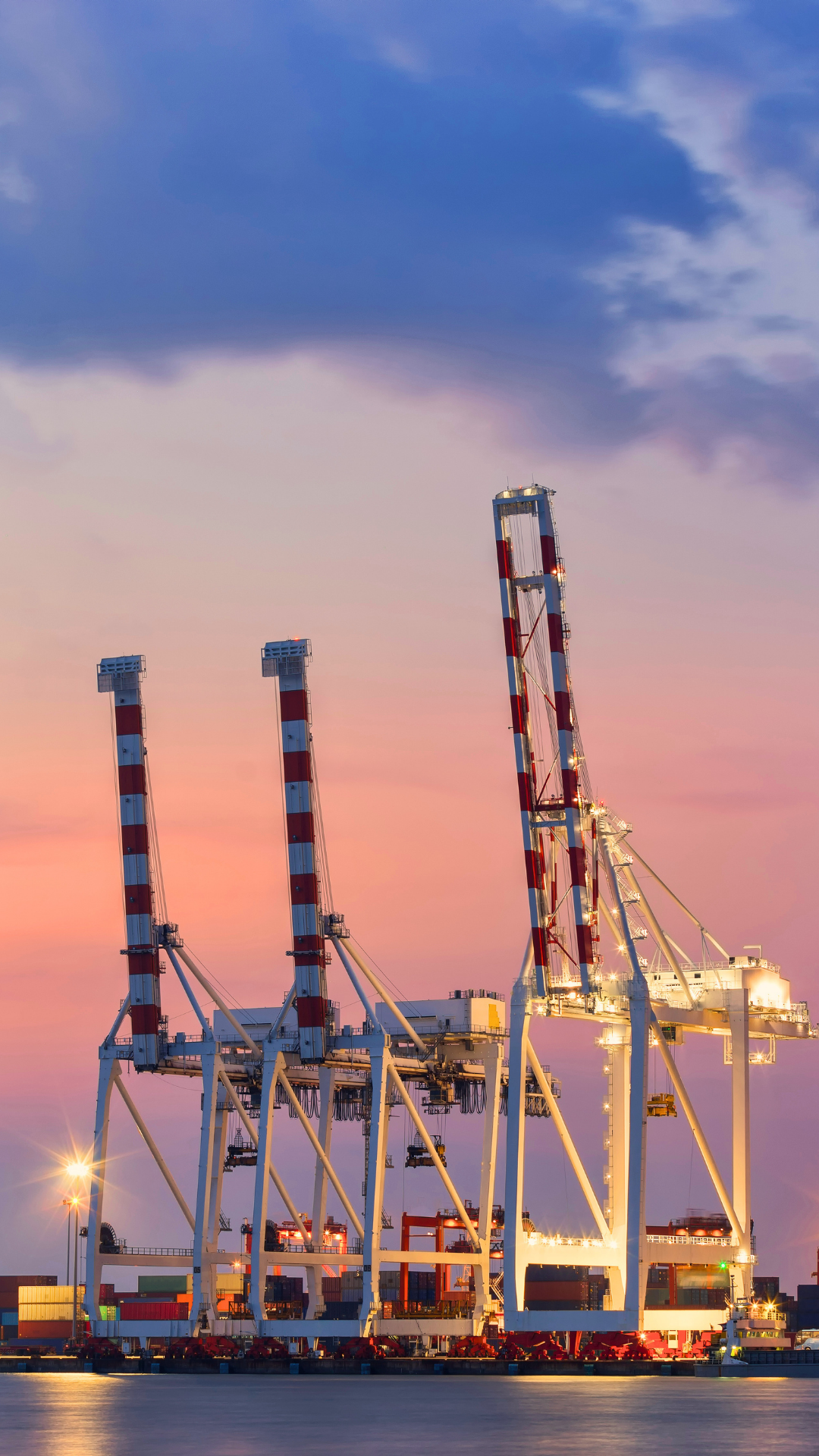 A port with large white and red striped container cranes at sunset, with a colorful sky and water in the foreground.